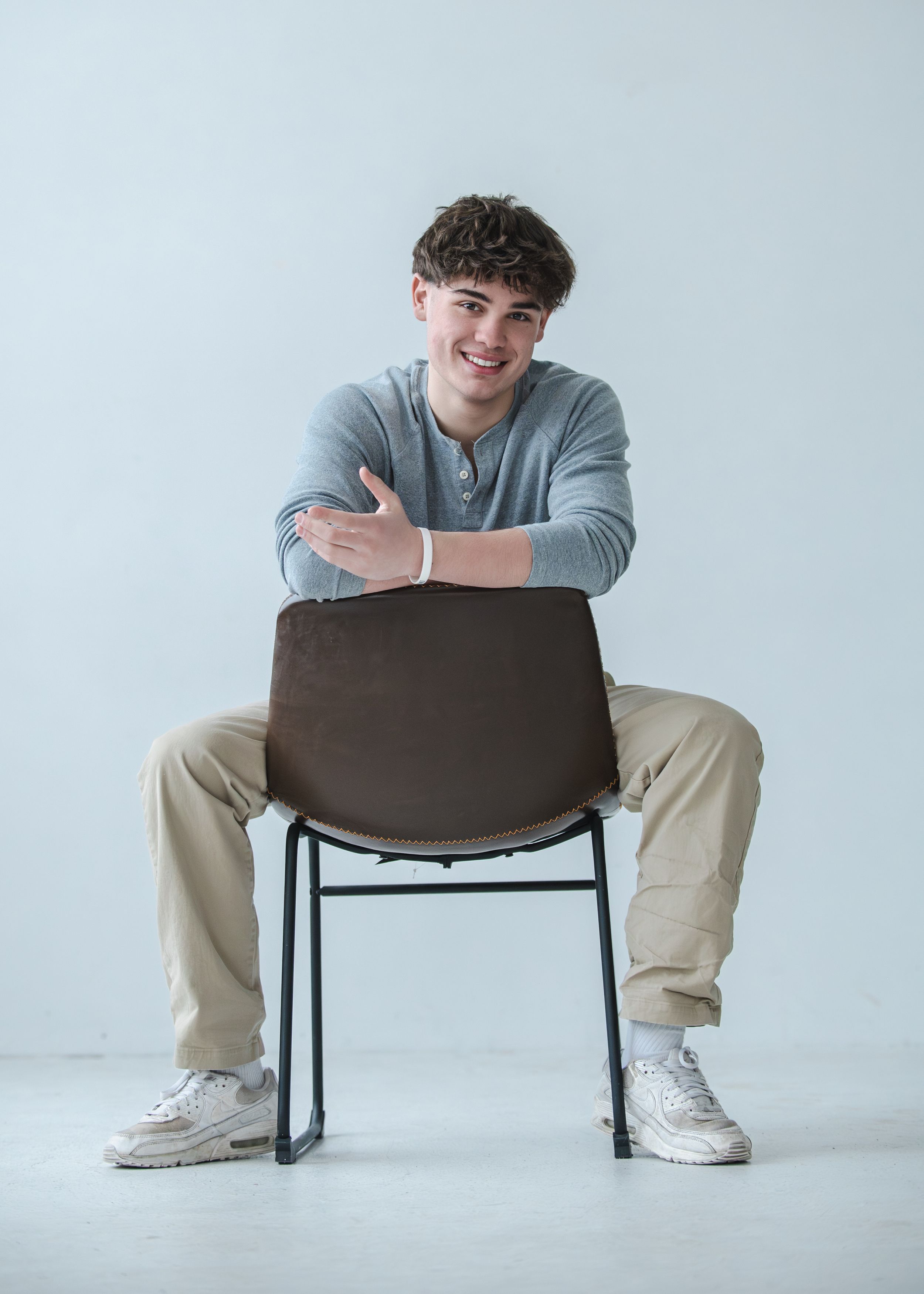 Young man smiling, sitting backwards on a brown chair, wearing a gray shirt and khaki pants in a studio setting.
