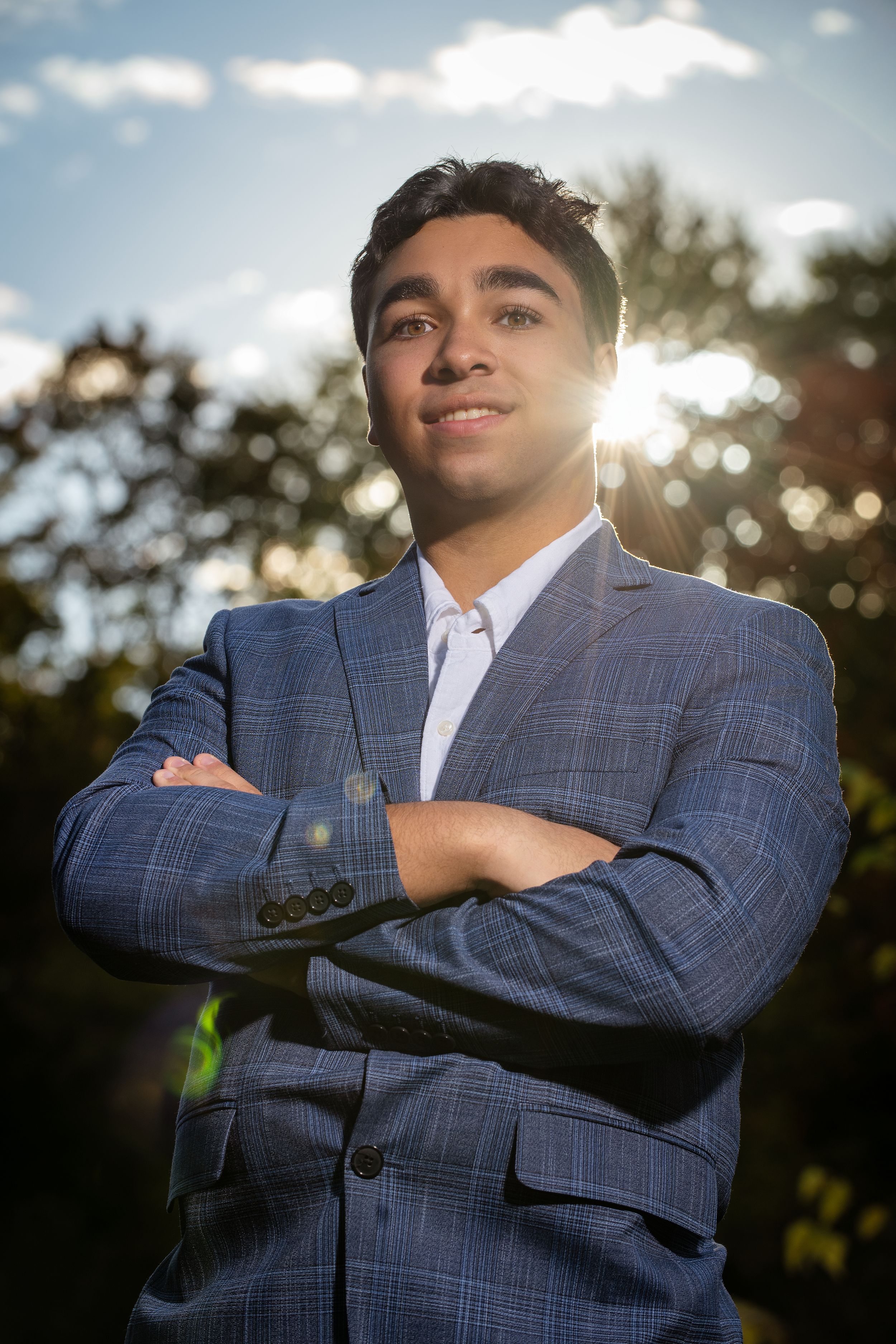 Young man in blue plaid suit stands confidently with arms crossed outdoors, backlit by sunlight through trees.