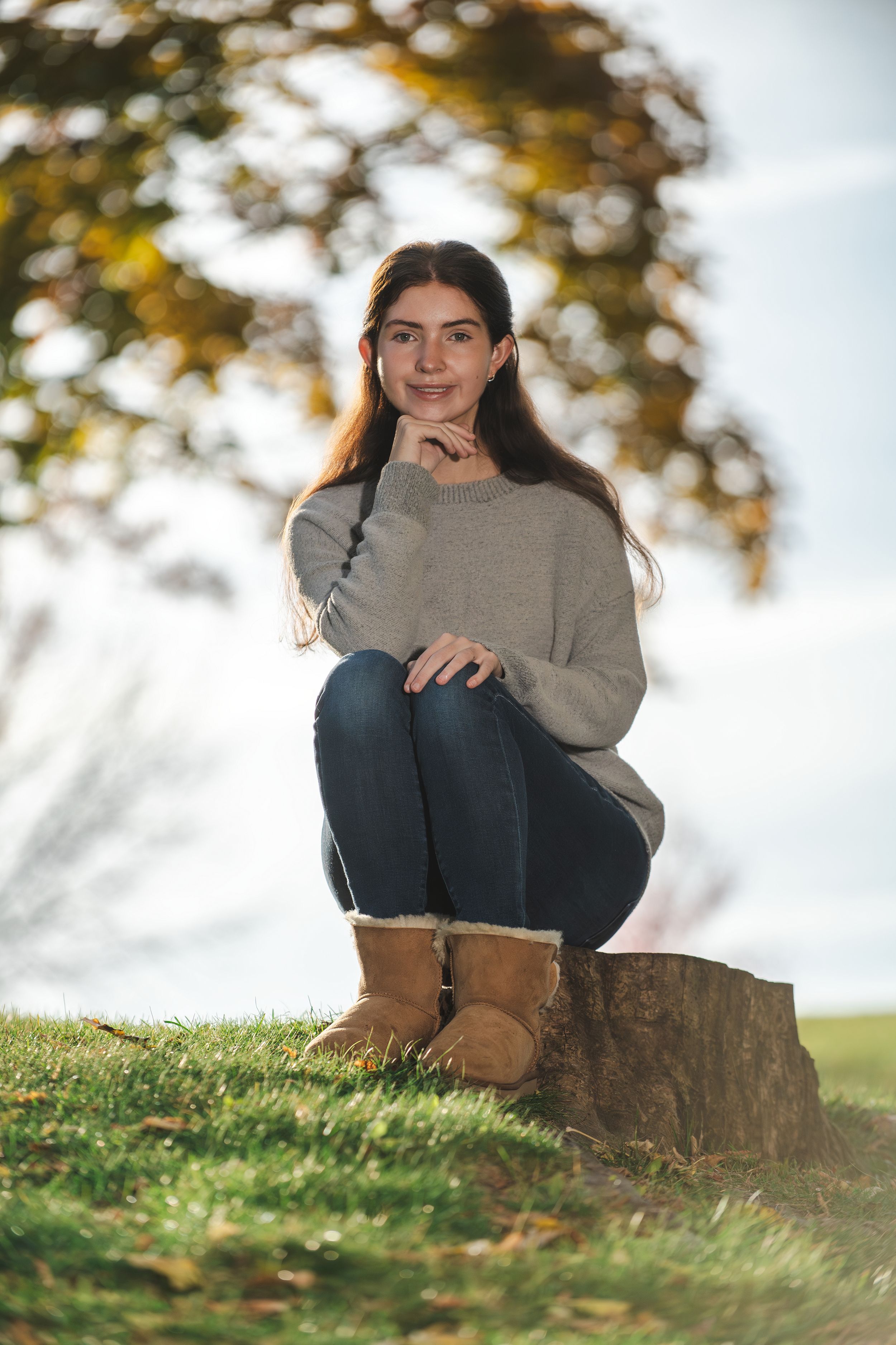 Young woman in gray sweater, jeans, and UGG boots sits on a tree stump in a park during autumn.