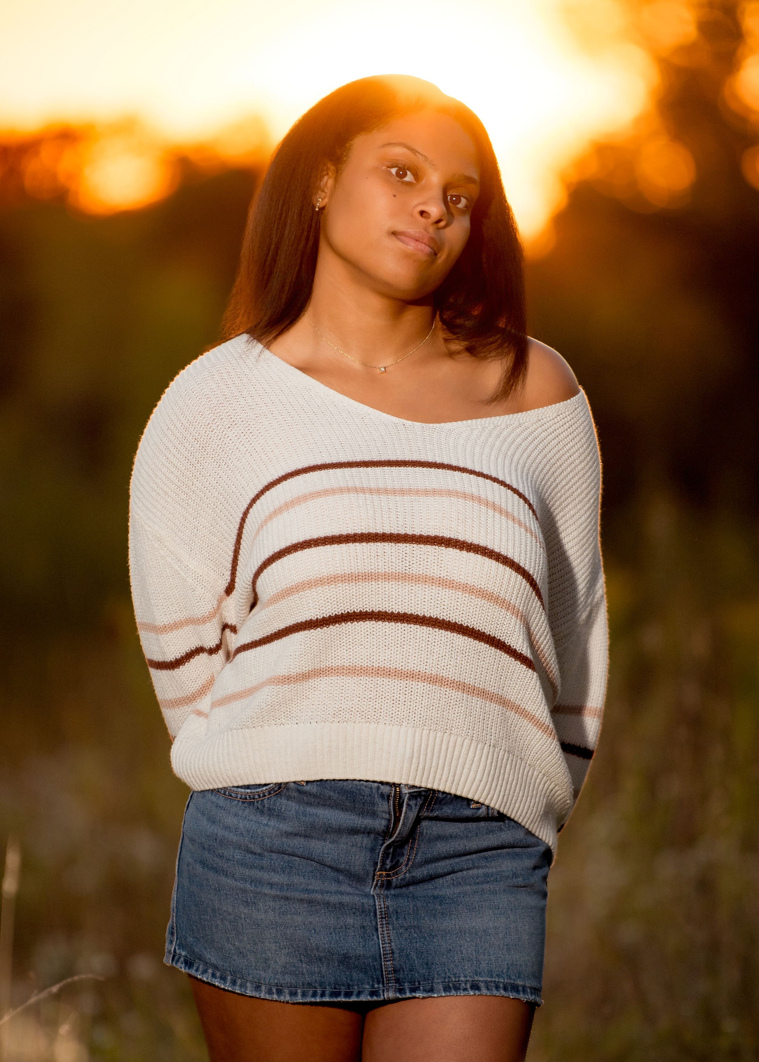 Young woman in striped sweater and denim skirt poses outdoors at golden hour with warm sunset light behind her.