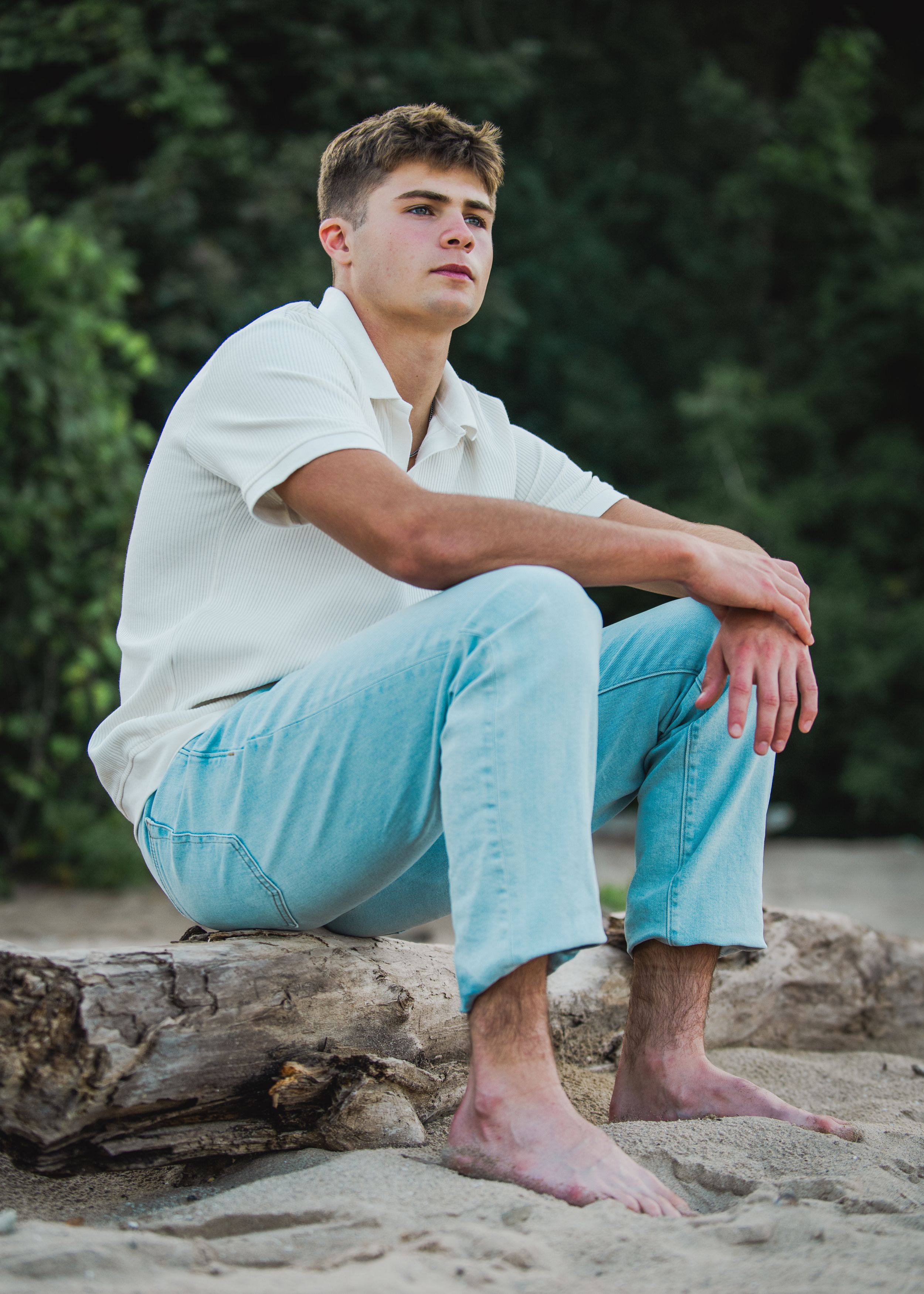 Young man in white shirt and light blue jeans sitting barefoot on driftwood at a sandy beach.