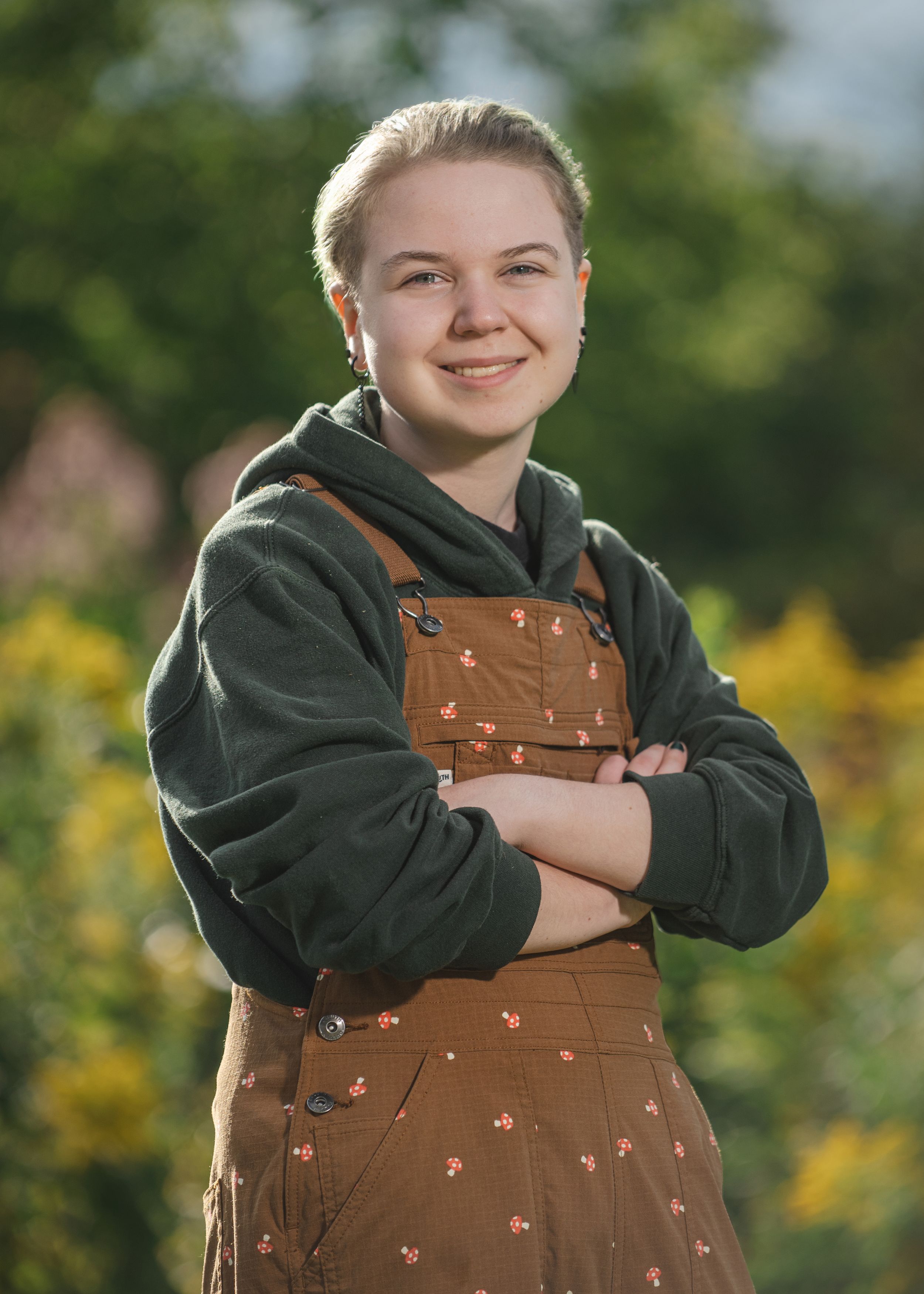 Smiling young person in brown floral overalls and green hoodie posing outdoors with arms crossed.