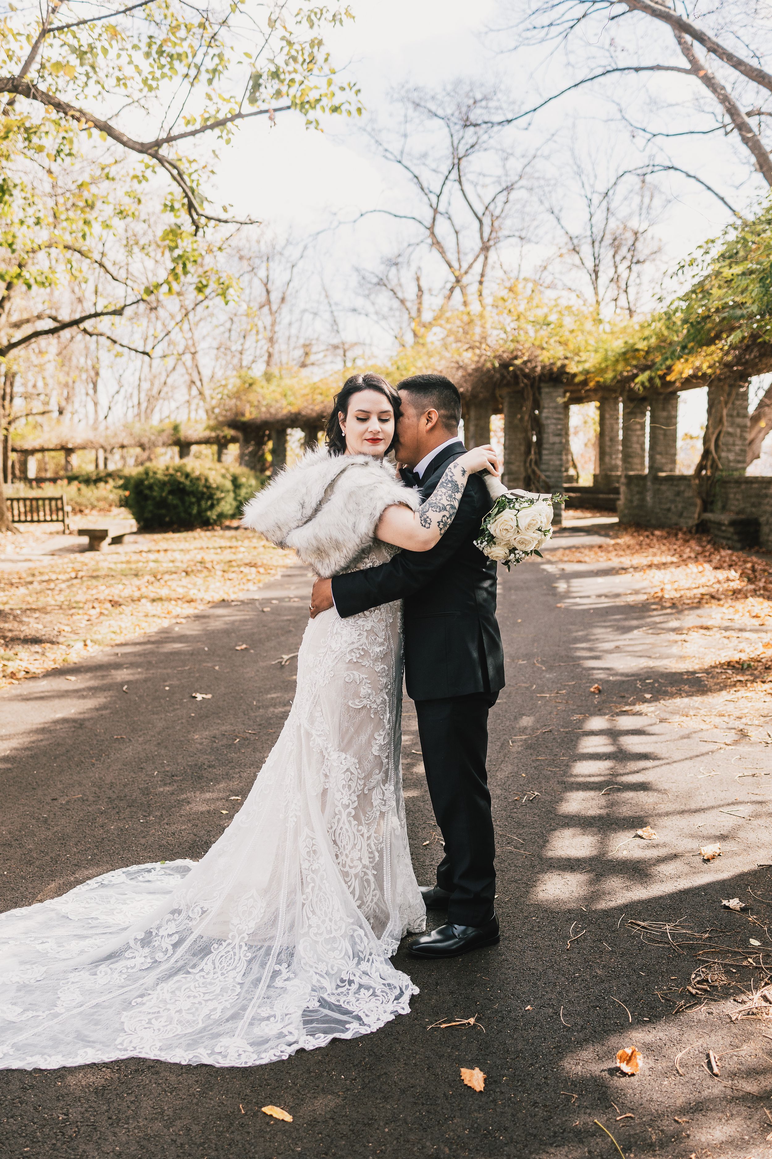 A bride in a lace gown with fur stole embraces her groom in a park during an autumn wedding portrait.