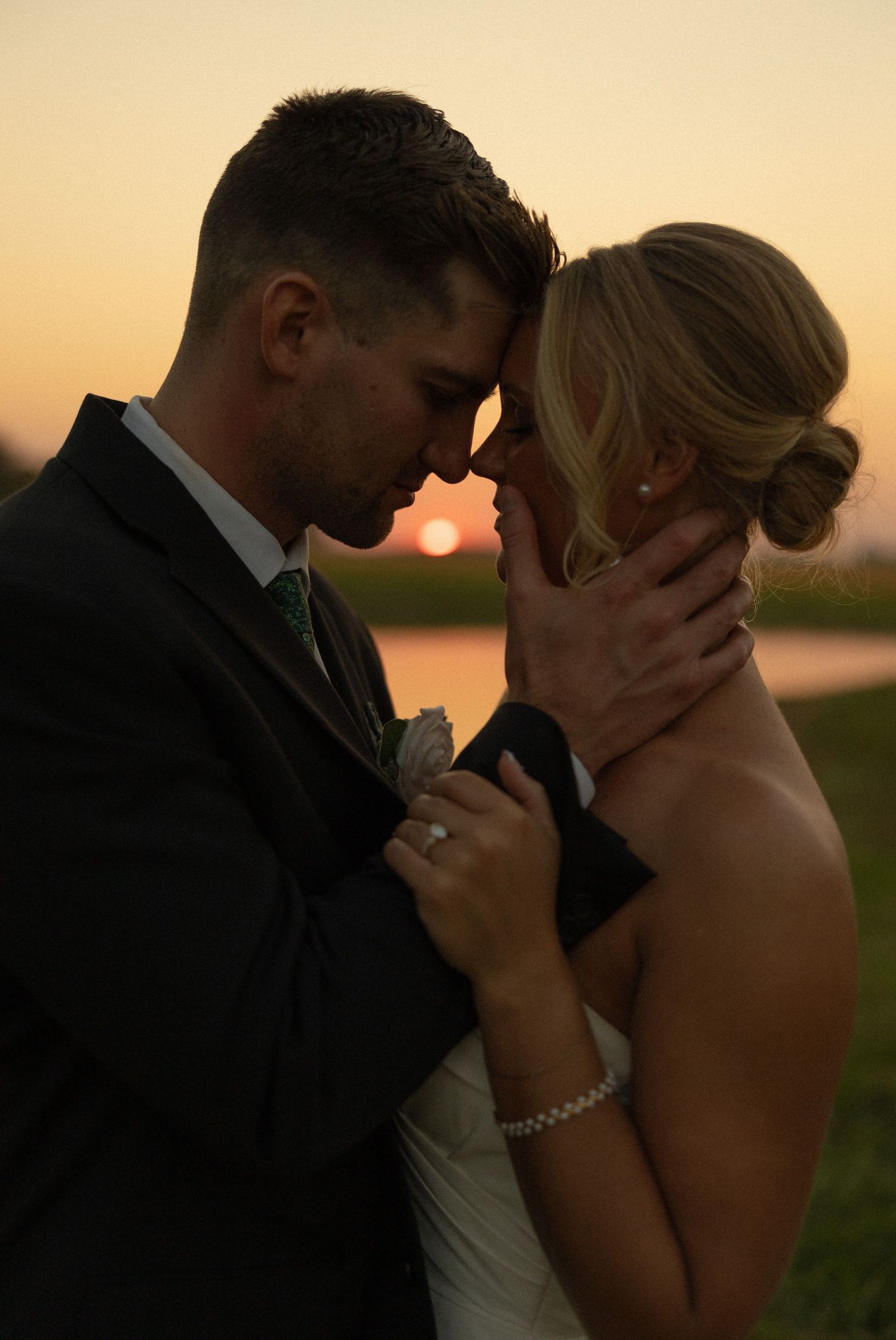 A romantic sunset photo of a couple embracing at a lakeside wedding ceremony in formal attire.