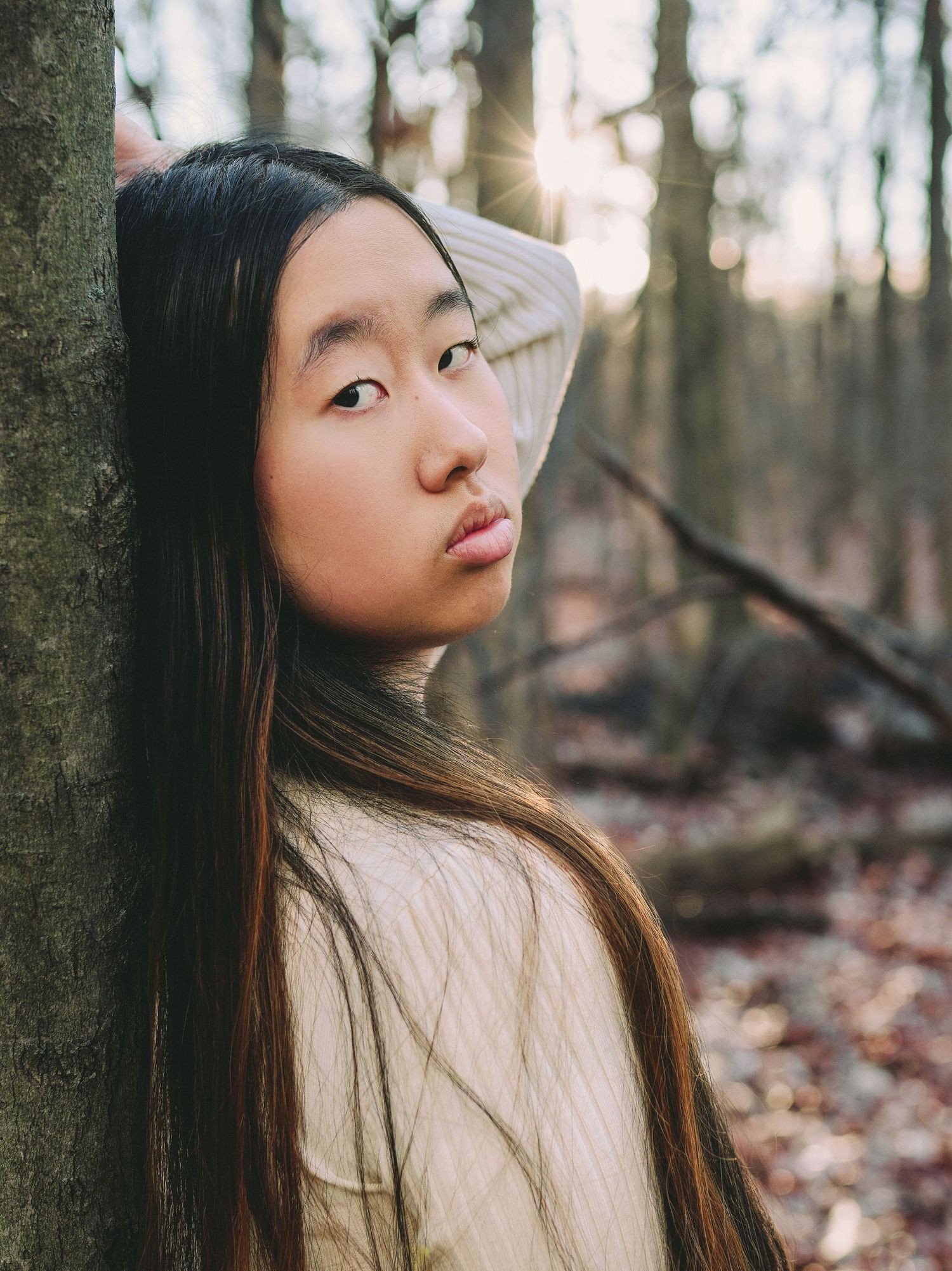 A serene forest portrait captures long dark hair and soft lighting through trees with fallen autumn leaves.