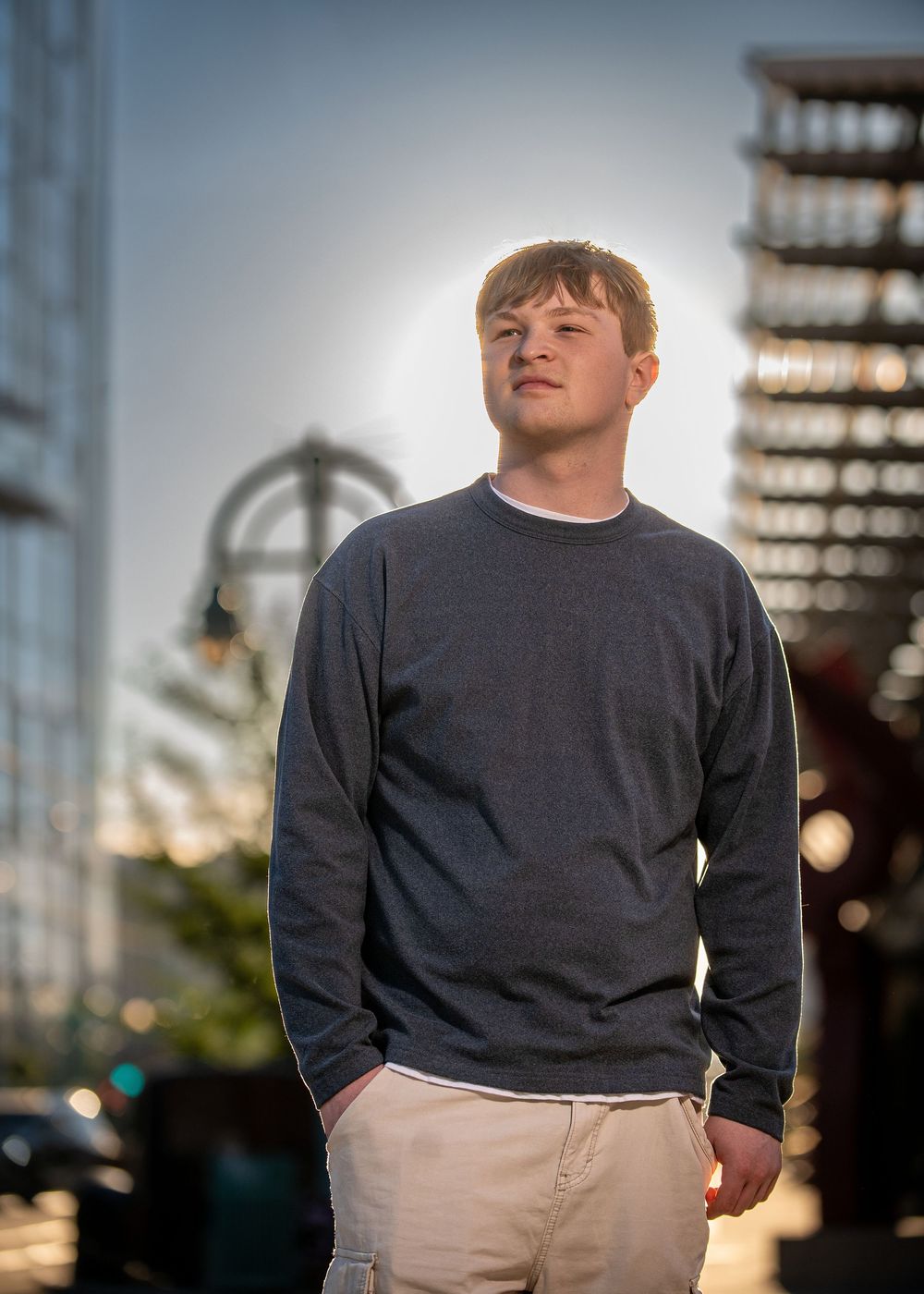 Young man in gray sweatshirt and khaki pants stands outdoors with sunlight behind him near industrial structures.