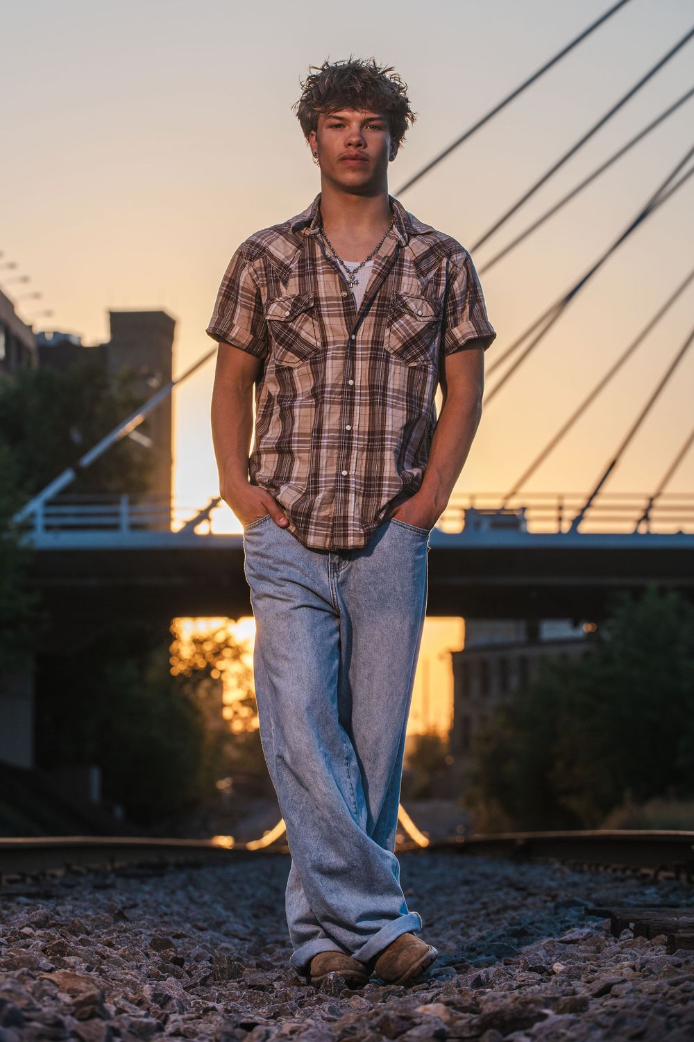 Young man in plaid shirt and jeans standing on railroad tracks at sunset with bridge in background.
