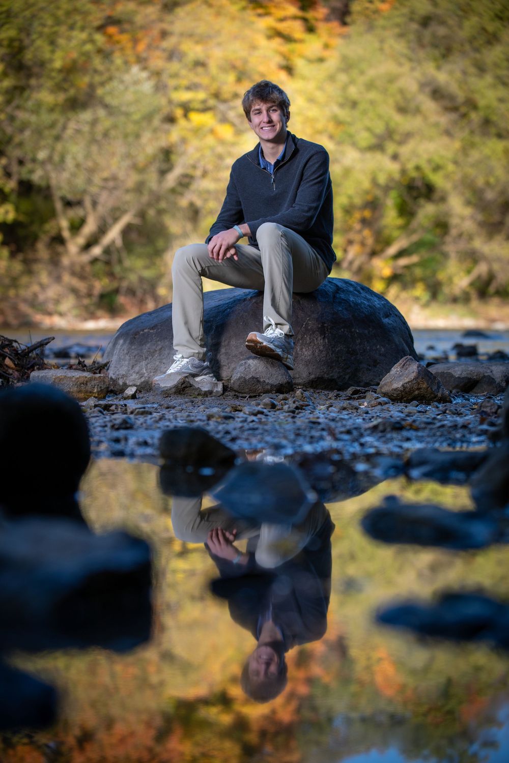 Man sitting on a rock by a stream with autumn foliage, his reflection visible in the calm water below.