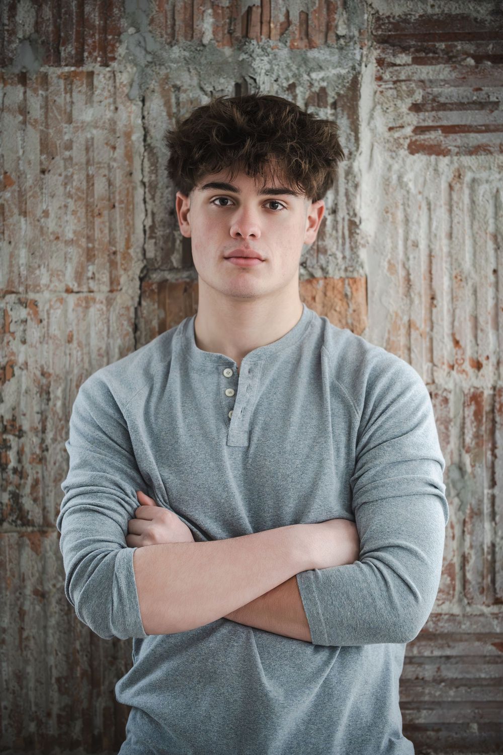 Young man with curly hair in gray henley shirt stands with arms crossed against rustic brick wall.