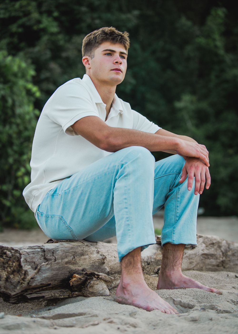 Young man in white shirt and light blue jeans sitting barefoot on driftwood at a sandy beach.