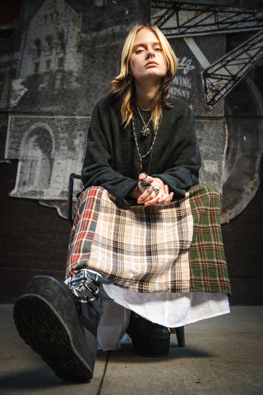Figure in black sweater and plaid skirt posed against industrial urban background.