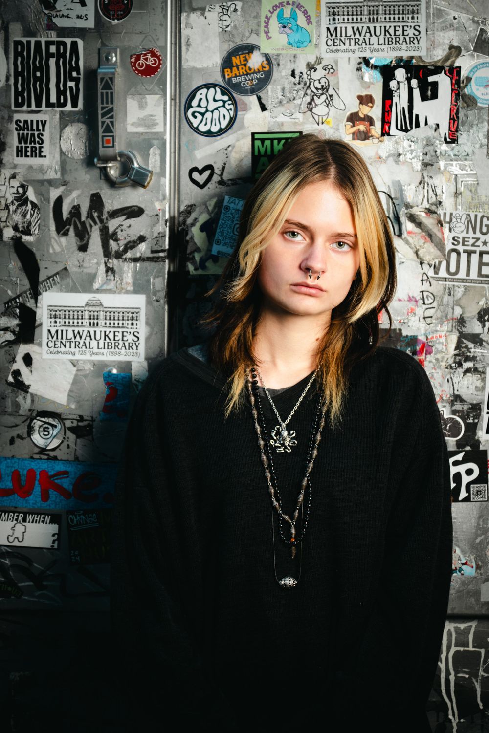 Portrait against wall covered in posters and graffiti featuring figure in black sweater and necklaces.