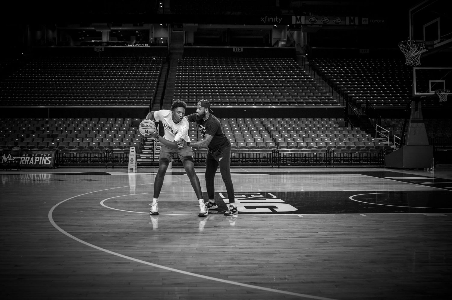 Two people practicing basketball moves on an empty court in black and white.