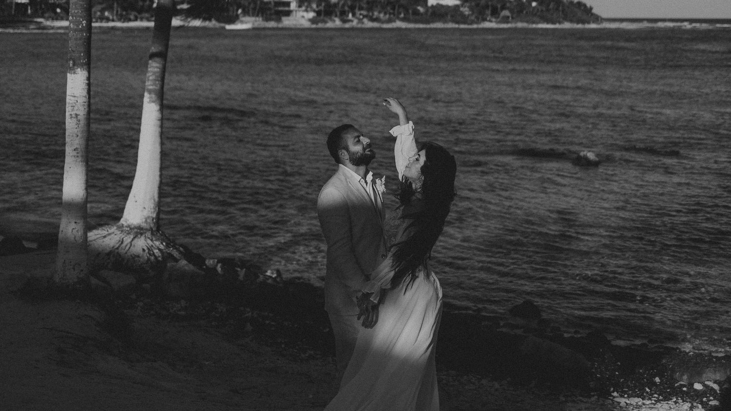 A romantic black and white moment captured between two people by white tree trunks near a waterfront.