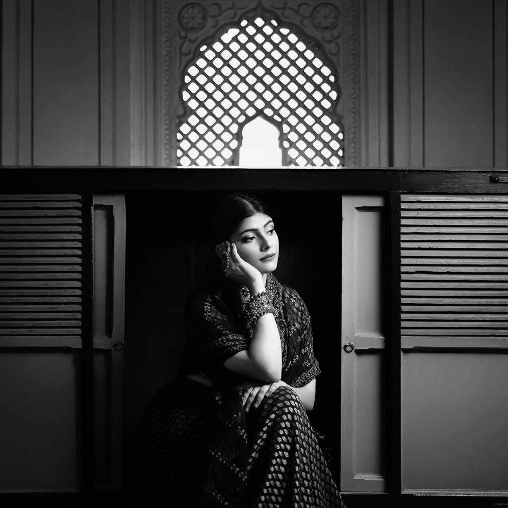 Black and white portrait of a lady in traditional attire seated beneath an ornate arched window with lattice pattern shot at Uniyara Jaipur.