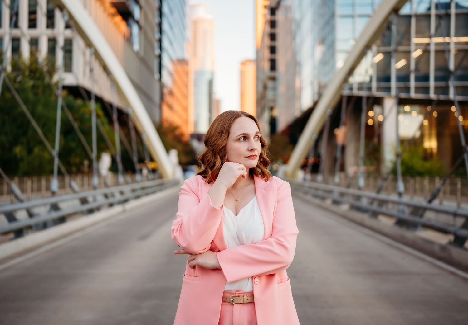 Woman in pink coat standing thoughtfully on urban bridge with city buildings in background at sunset.