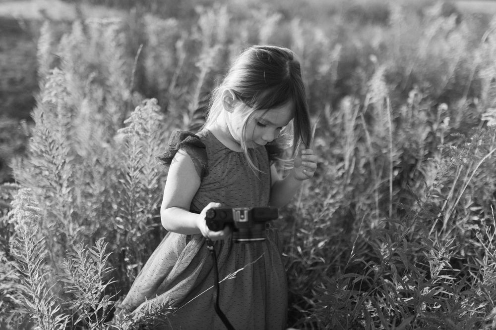 A black and white photo of a child in a dress exploring tall wildflowers in a field.