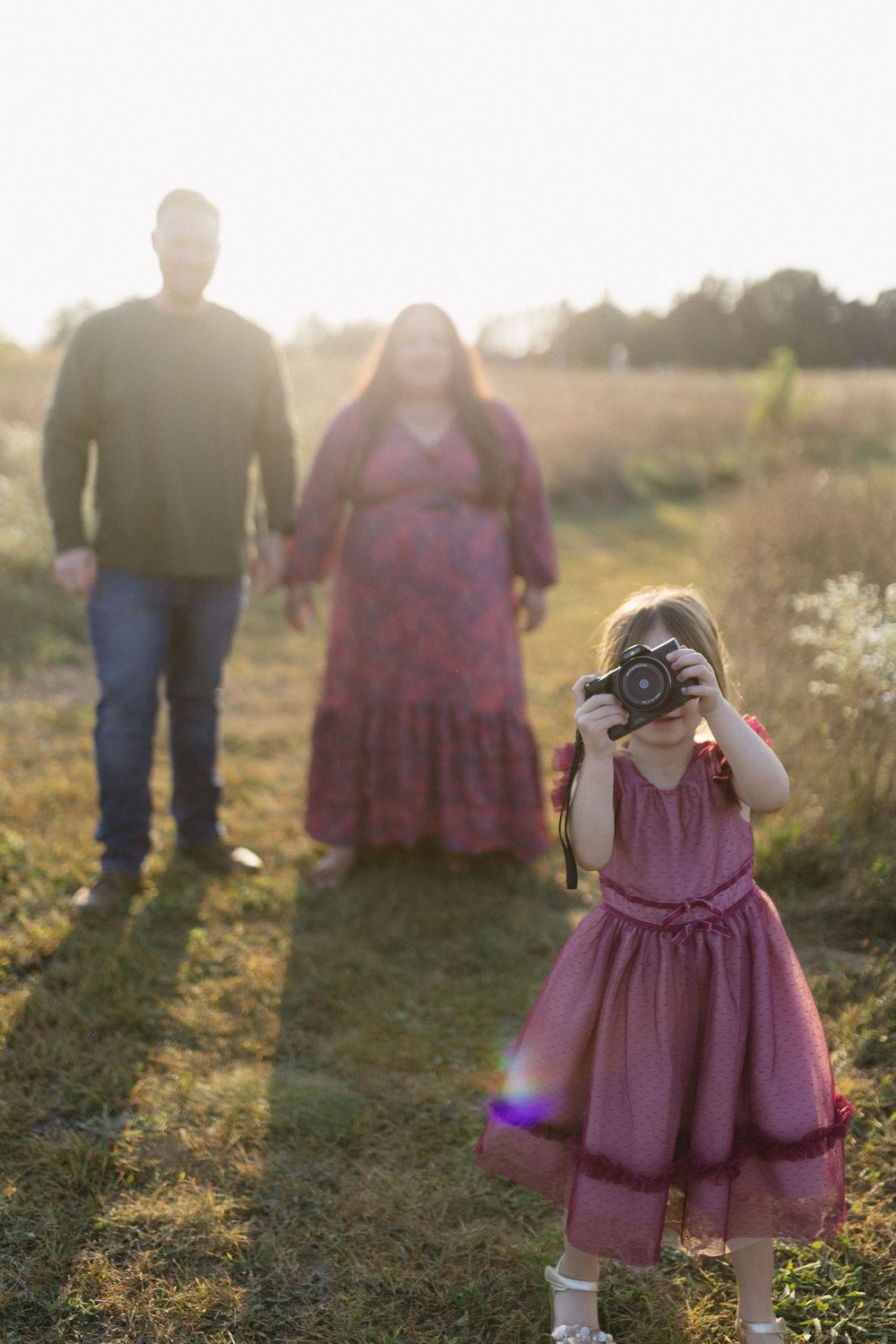 A young photographer in a pink dress takes pictures while adults watch in golden sunset light.