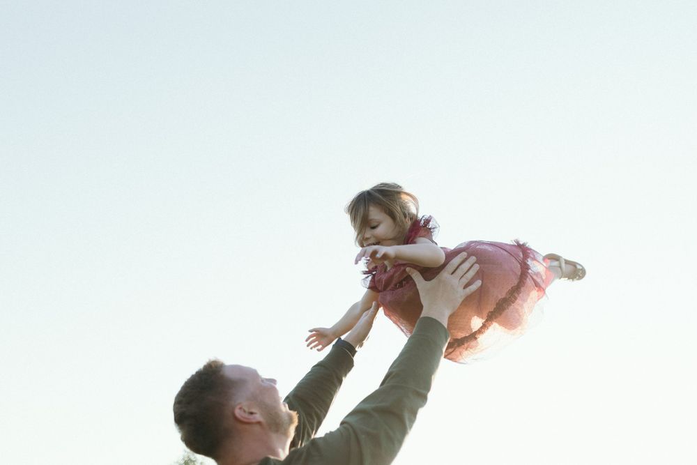 A silhouetted figure lifts a small child into the air against a bright sky.