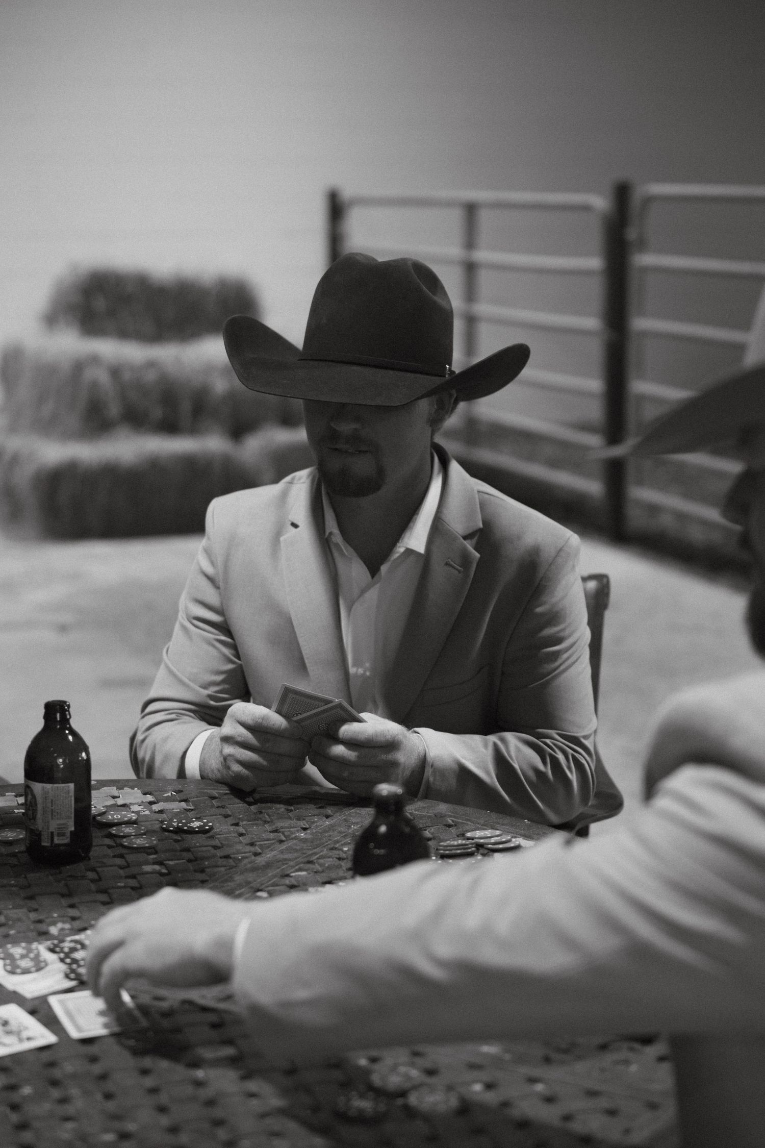 Man in cowboy hat and blazer playing cards at rustic table in black and white photo.