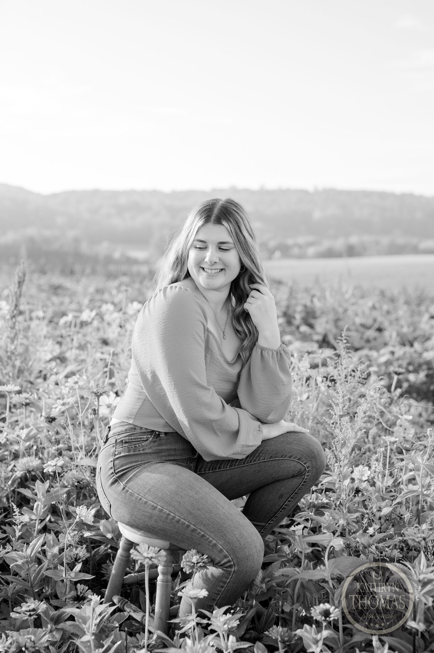 A black and white portrait of someone sitting on a stool in a field wearing jeans and a sweater.