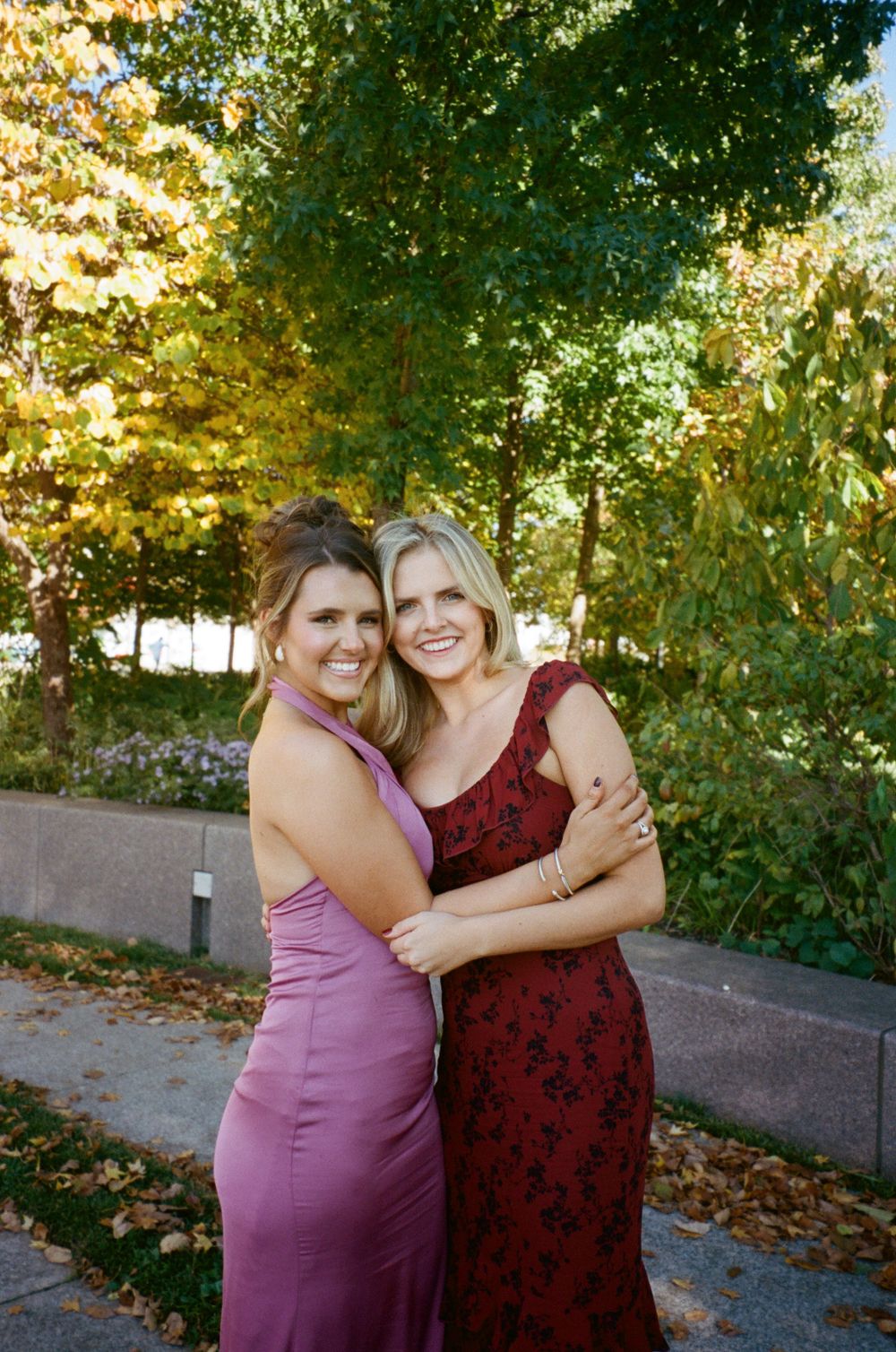 Two women in pink and burgundy formal dresses embracing outdoors.