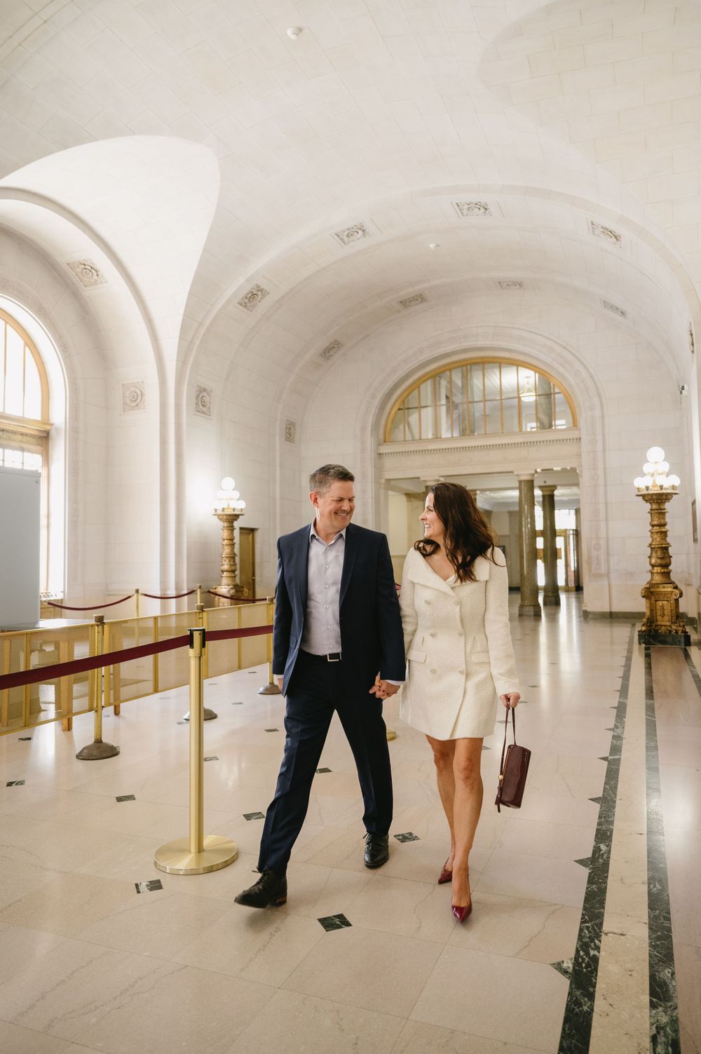 Couple walking through a grand marble hallway with arched ceilings and golden fixtures.