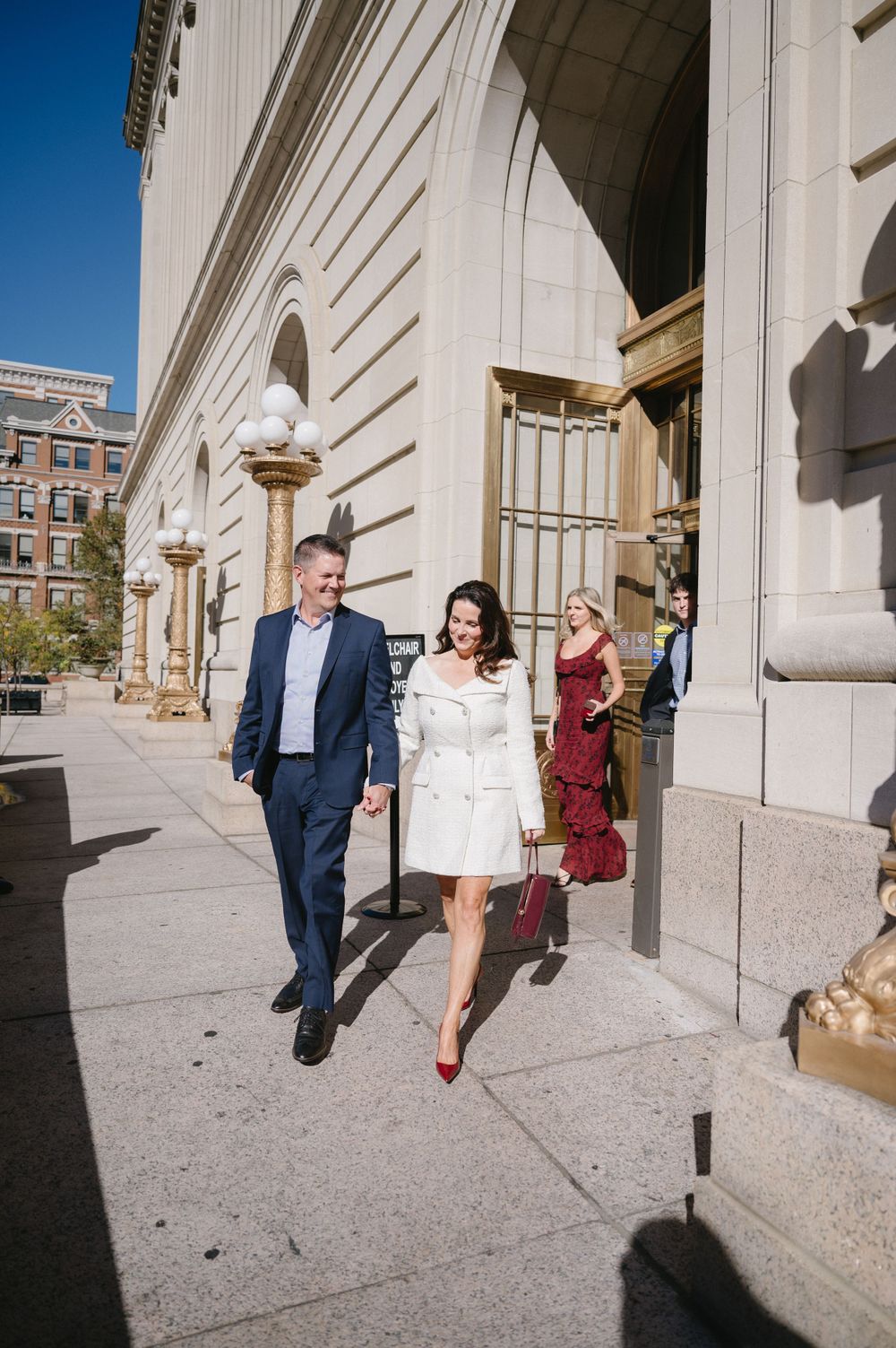 Well-dressed pair walking along a classical building exterior on a sunny day.