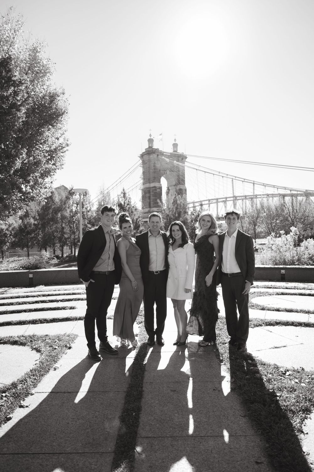 Black and white photo of group standing in front of suspension bridge with sun flare.