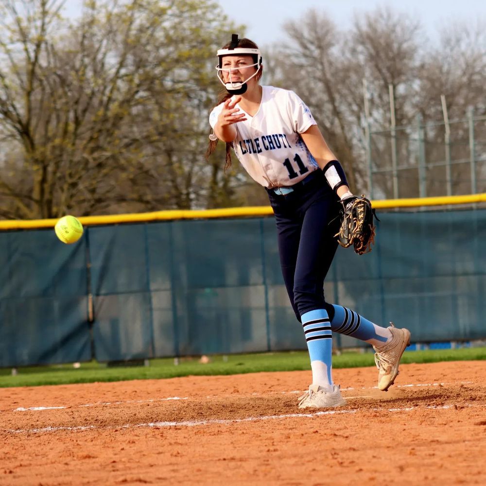 Softball pitcher in white uniform delivers pitch on dirt mound during game.