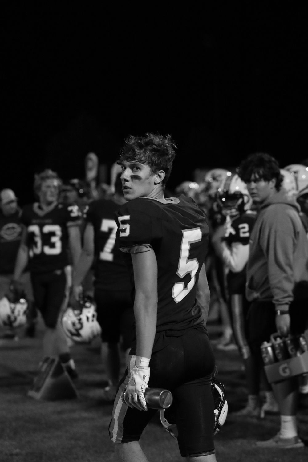Black and white photo of football player in dark uniform standing on sideline.