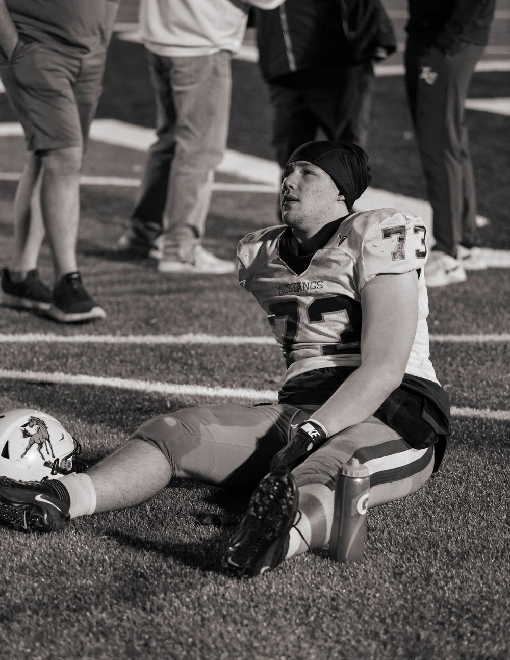 Football player in black beanie rests on sideline during game.