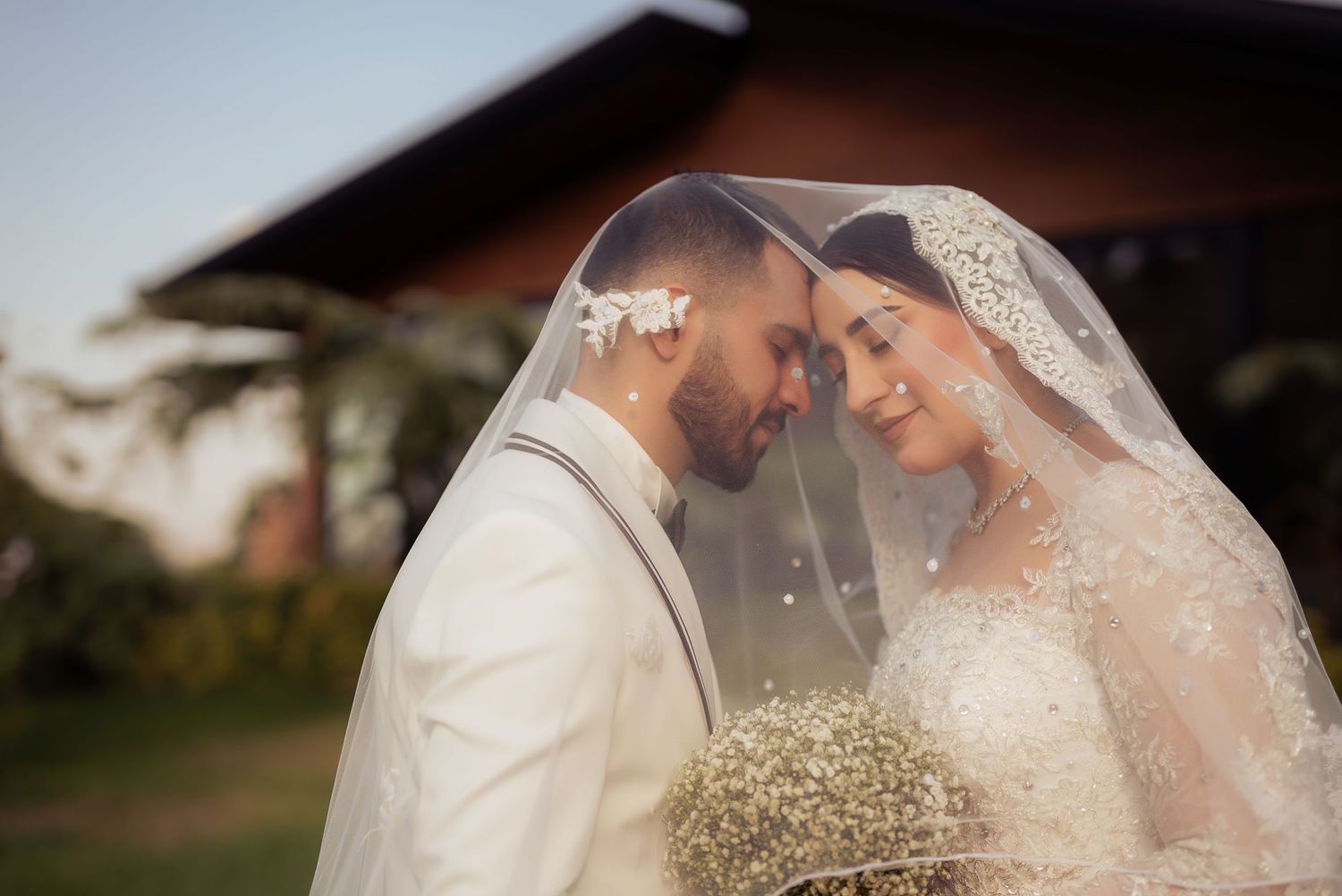 A romantic wedding couple shares an intimate moment under a delicate lace veil while holding baby's breath flowers.