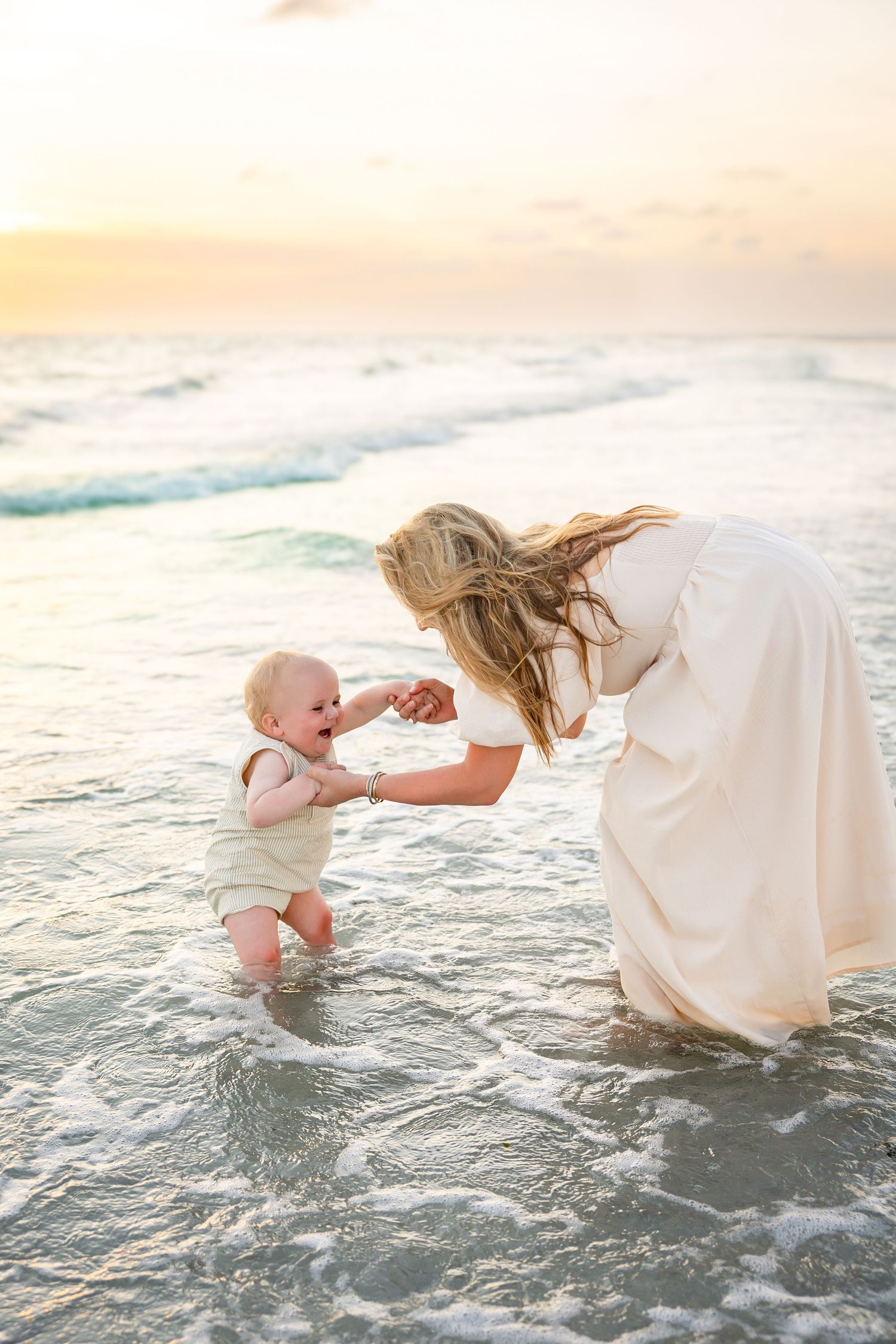 Mother in white dress helping smiling baby take first steps in ocean waves at sunset beach.