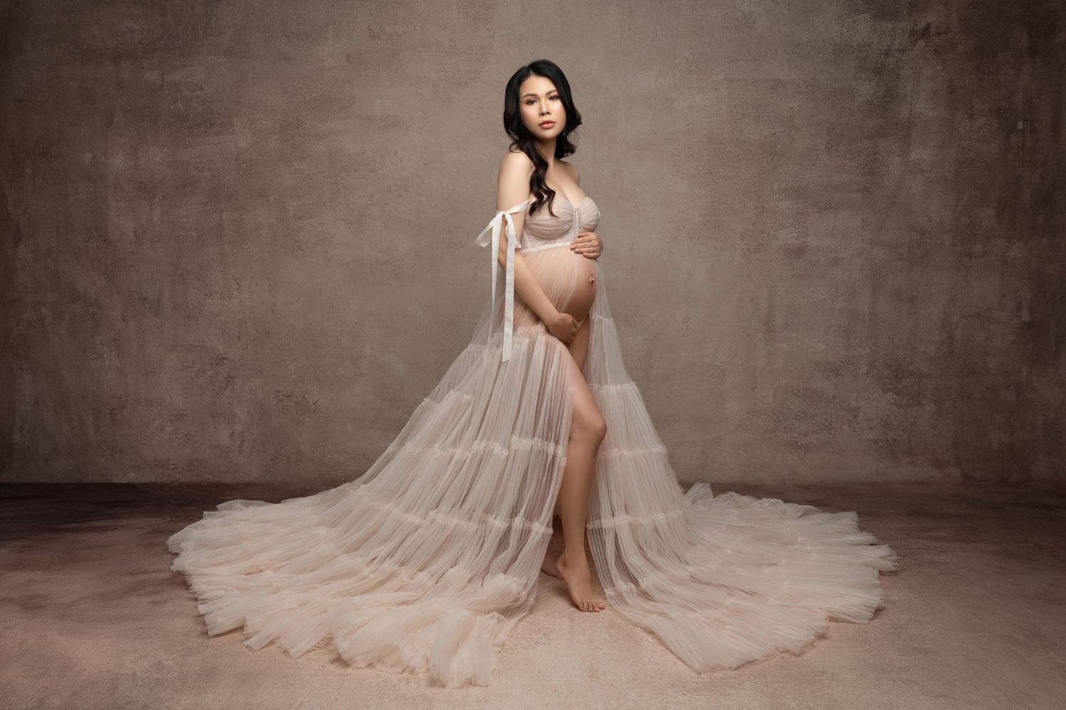 An elegant maternity photo featuring a sheer flowing gown against a textured brown backdrop in a studio setting.