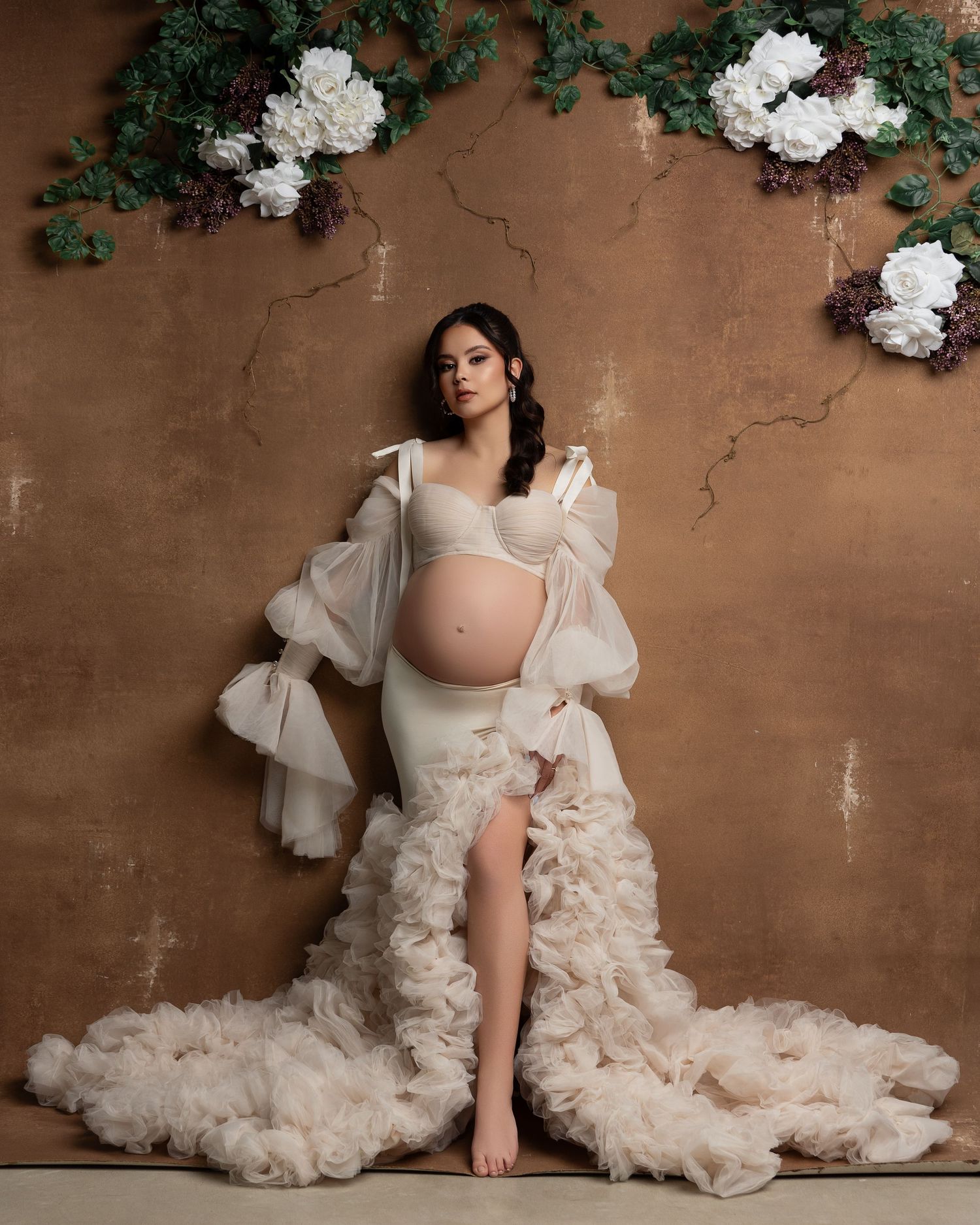 An elegant maternity portrait featuring white flowing fabrics and florals against a textured brown wall background.