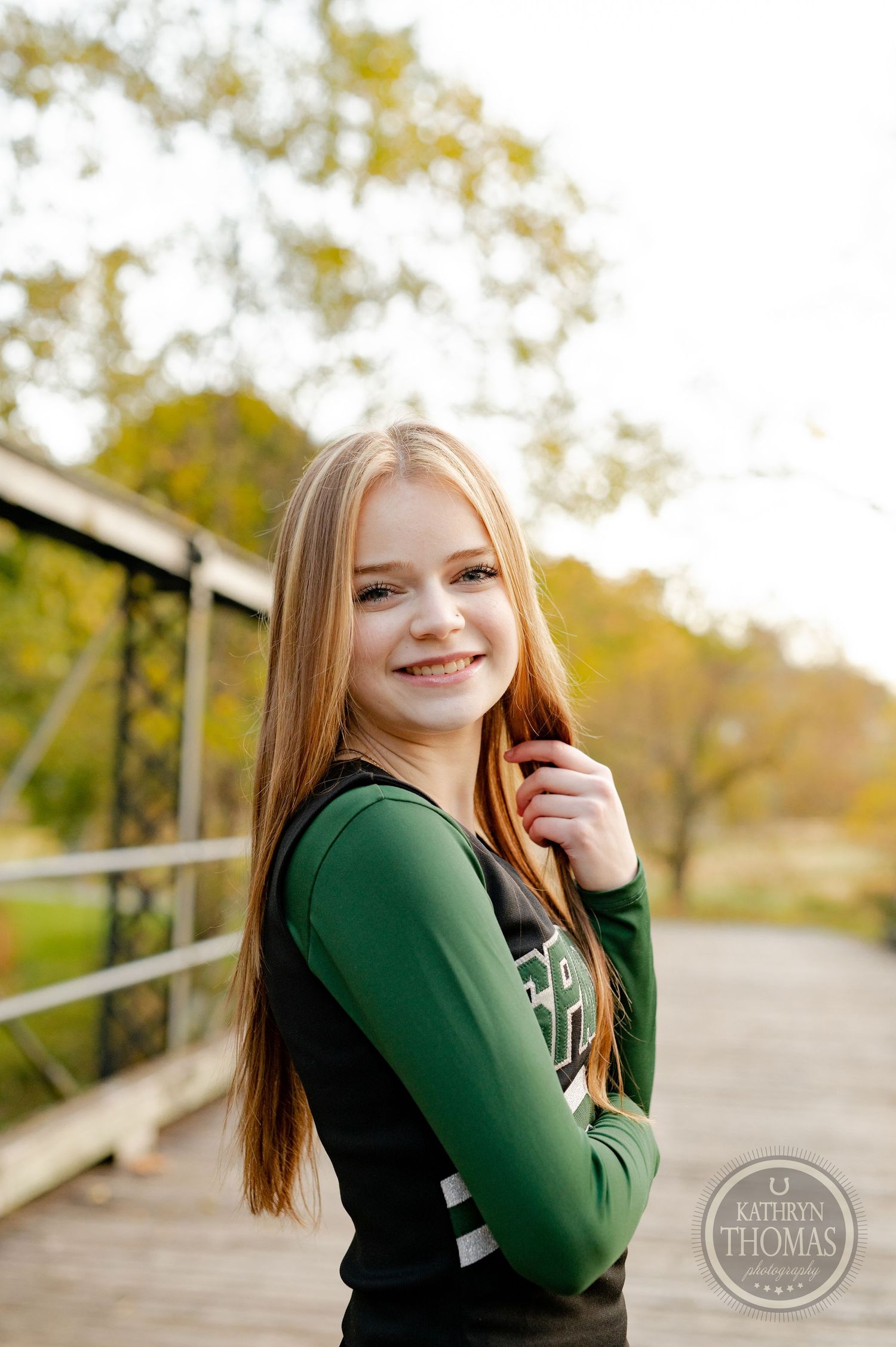 Cheerleader in green and black uniform poses against autumn foliage on wooden bridge.