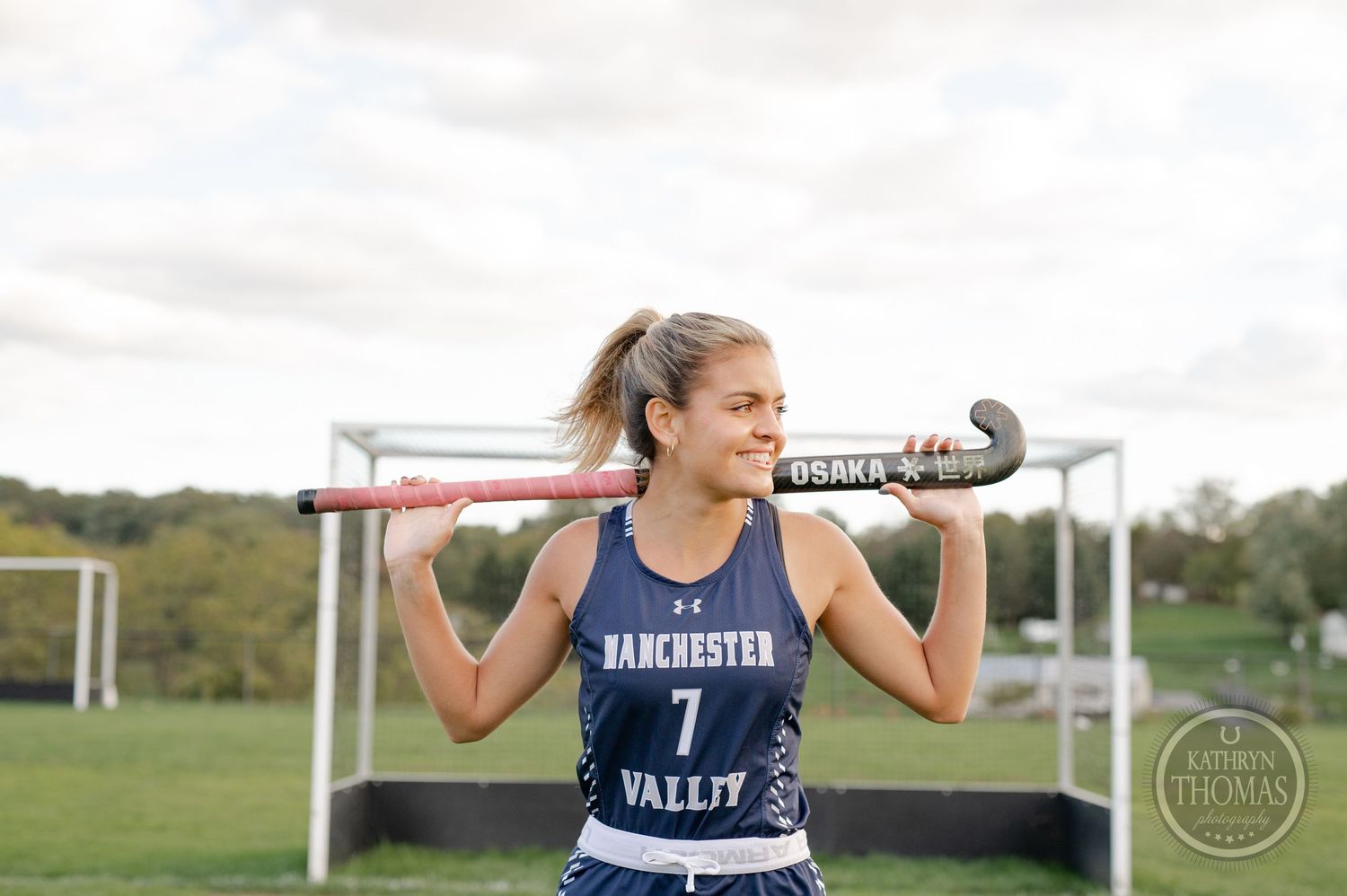 Field hockey athlete in blue uniform holding stick across shoulders with goal in background.