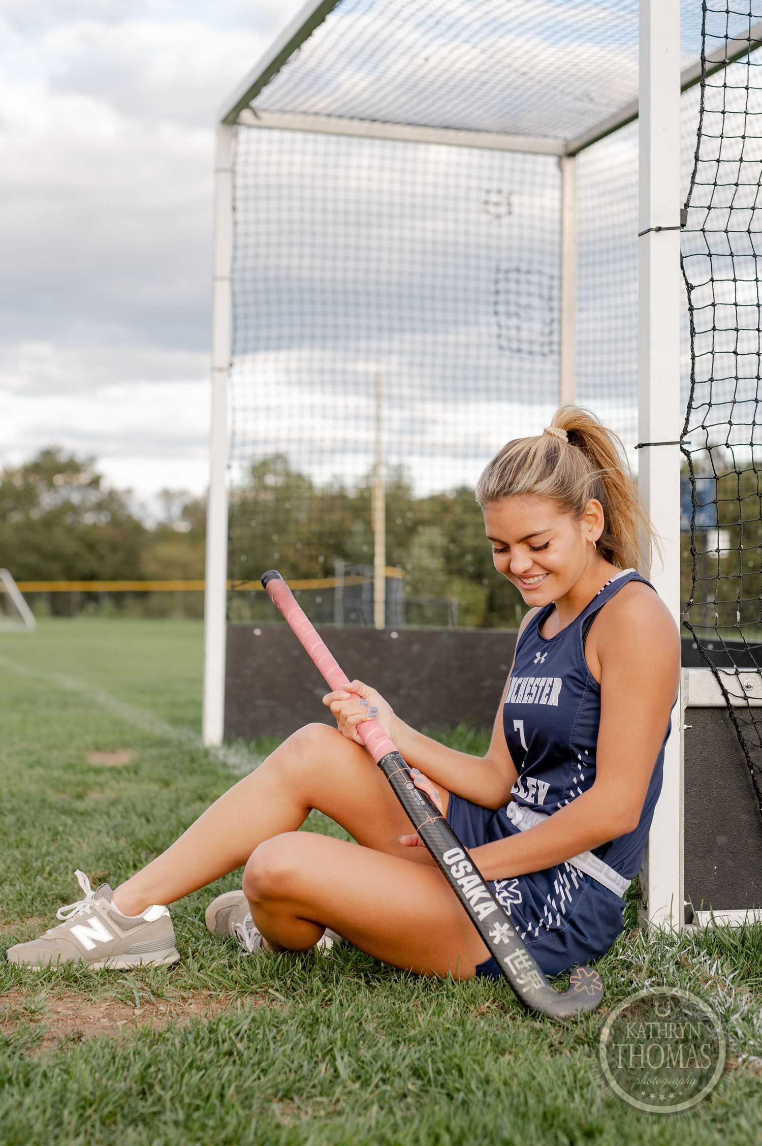 Field hockey player sits by goal post examining pink stick on grassy field.