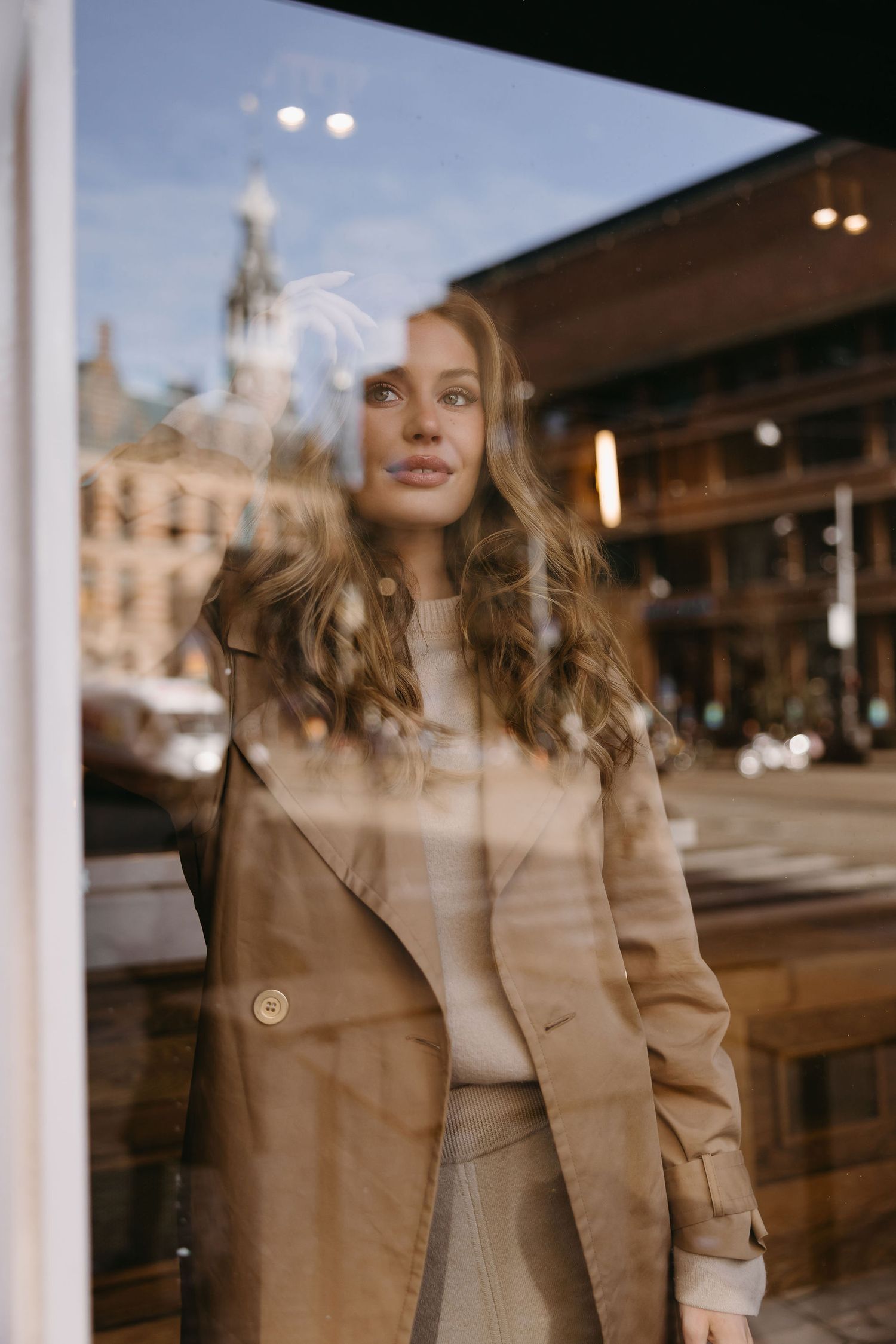 Stylish woman in beige coat standing by window with city reflection.