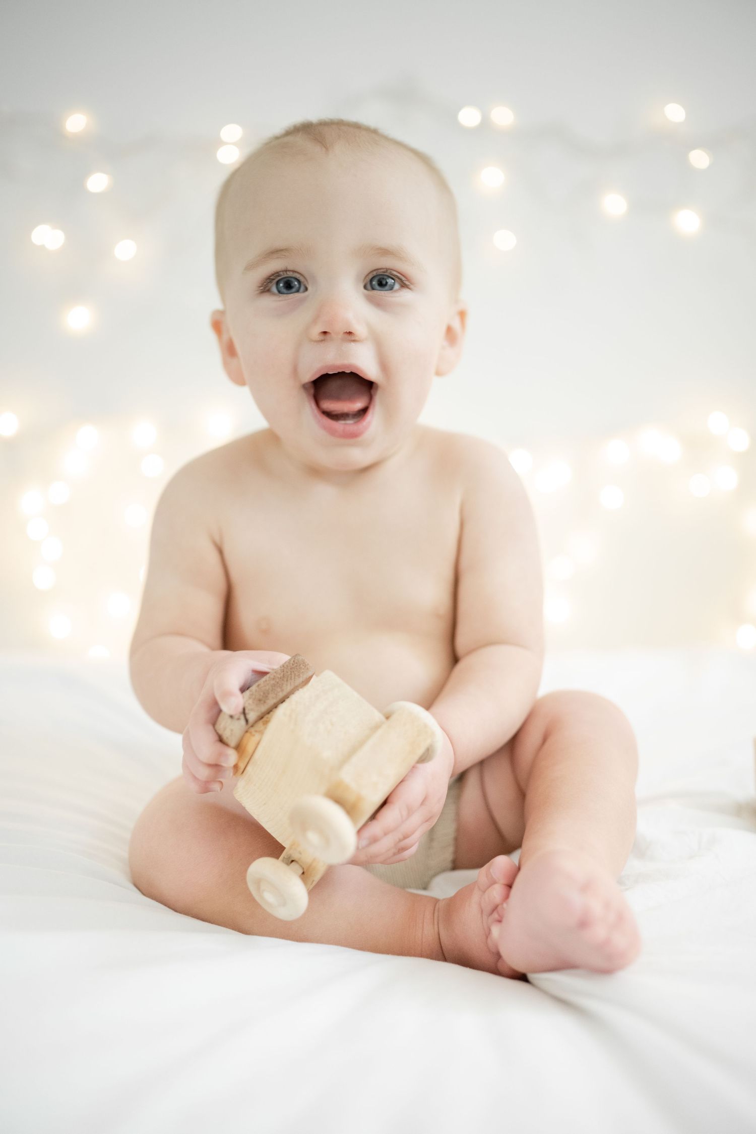 little girl smiles lovingly at the camera with a wooden toy car in her hands.  sitting on white bedding, twinkling light