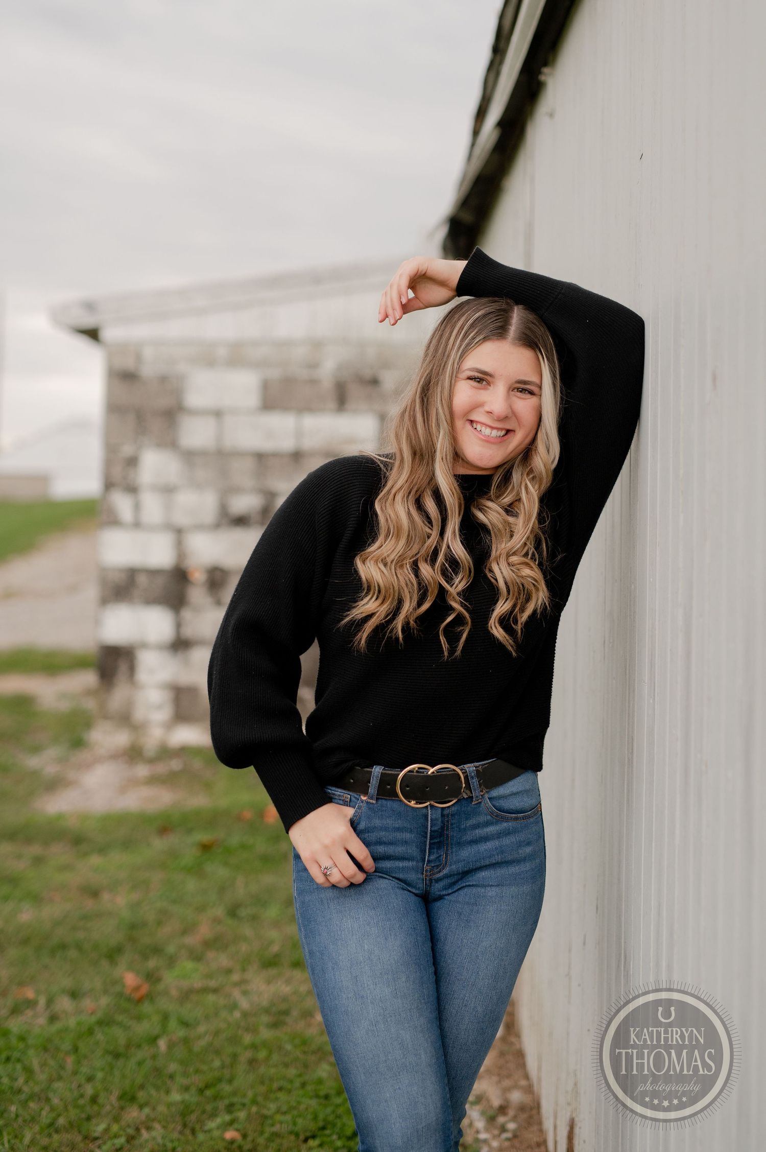 Playful outdoor portrait in black sweater and jeans against rustic white wall and stone foundation.