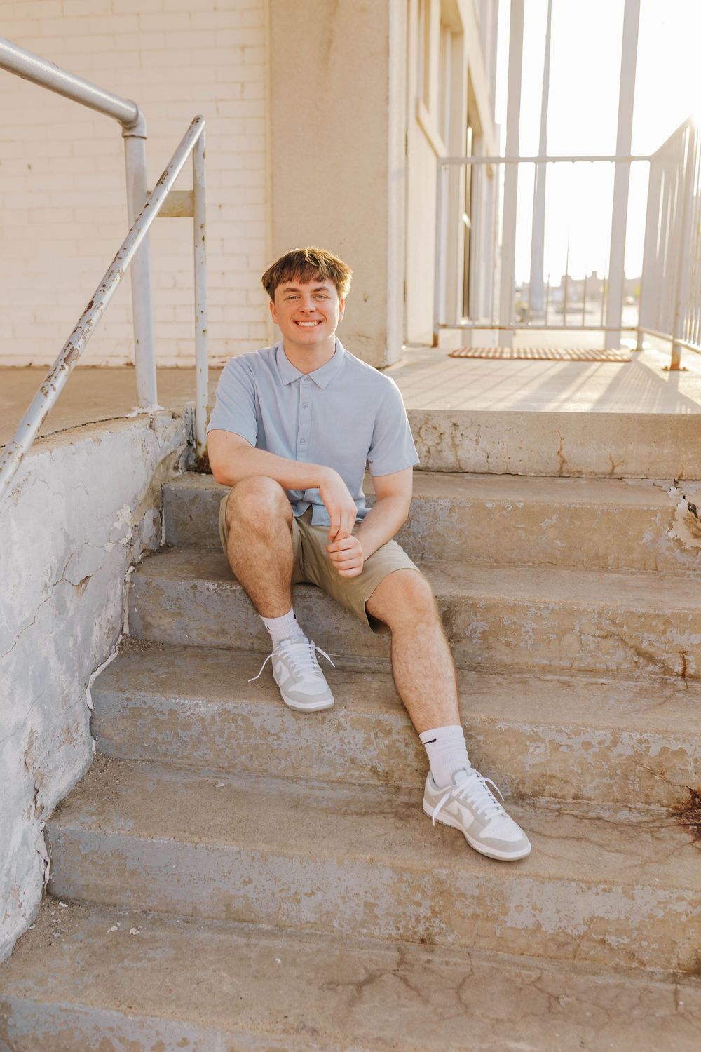 A person in a light blue polo shirt and khaki shorts sits casually on concrete steps in a sunlit space.