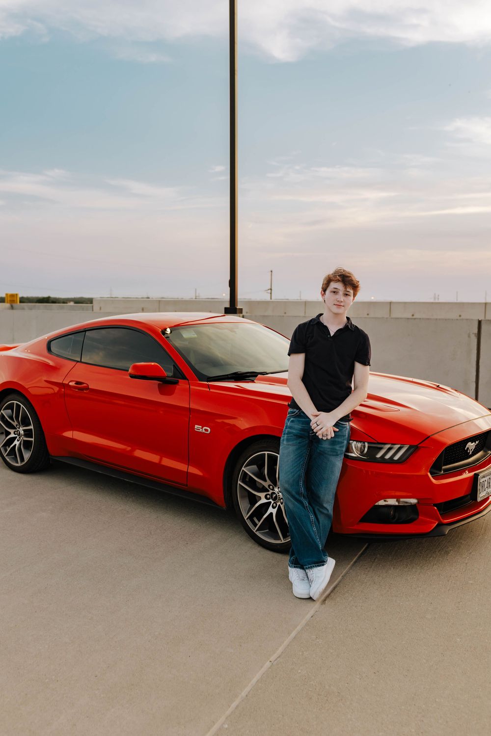 A bright red Ford Mustang GT parked on a rooftop parking garage with someone casually leaning against it.