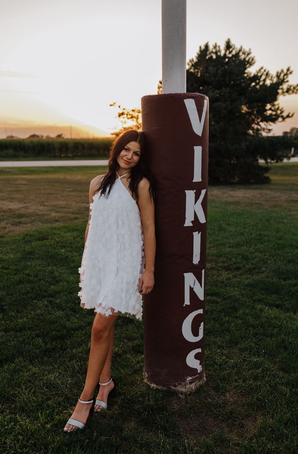 A portrait of someone in a white dress posed next to a Vikings sign pole at sunset.