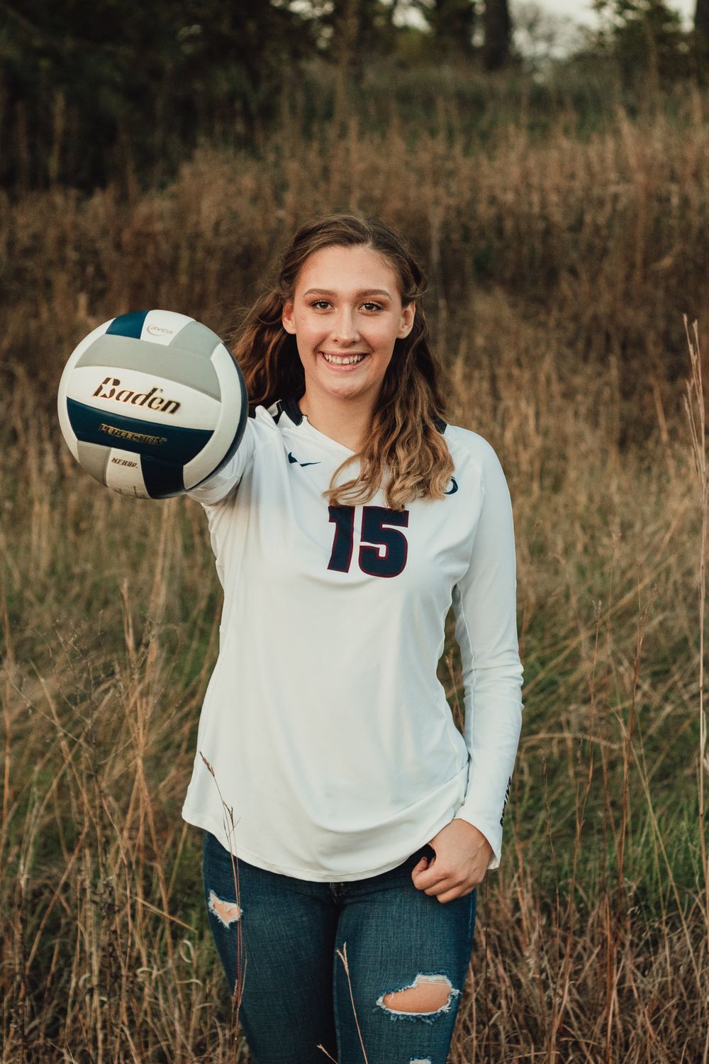An athlete poses with a volleyball wearing jersey number 15 in a rustic field setting.