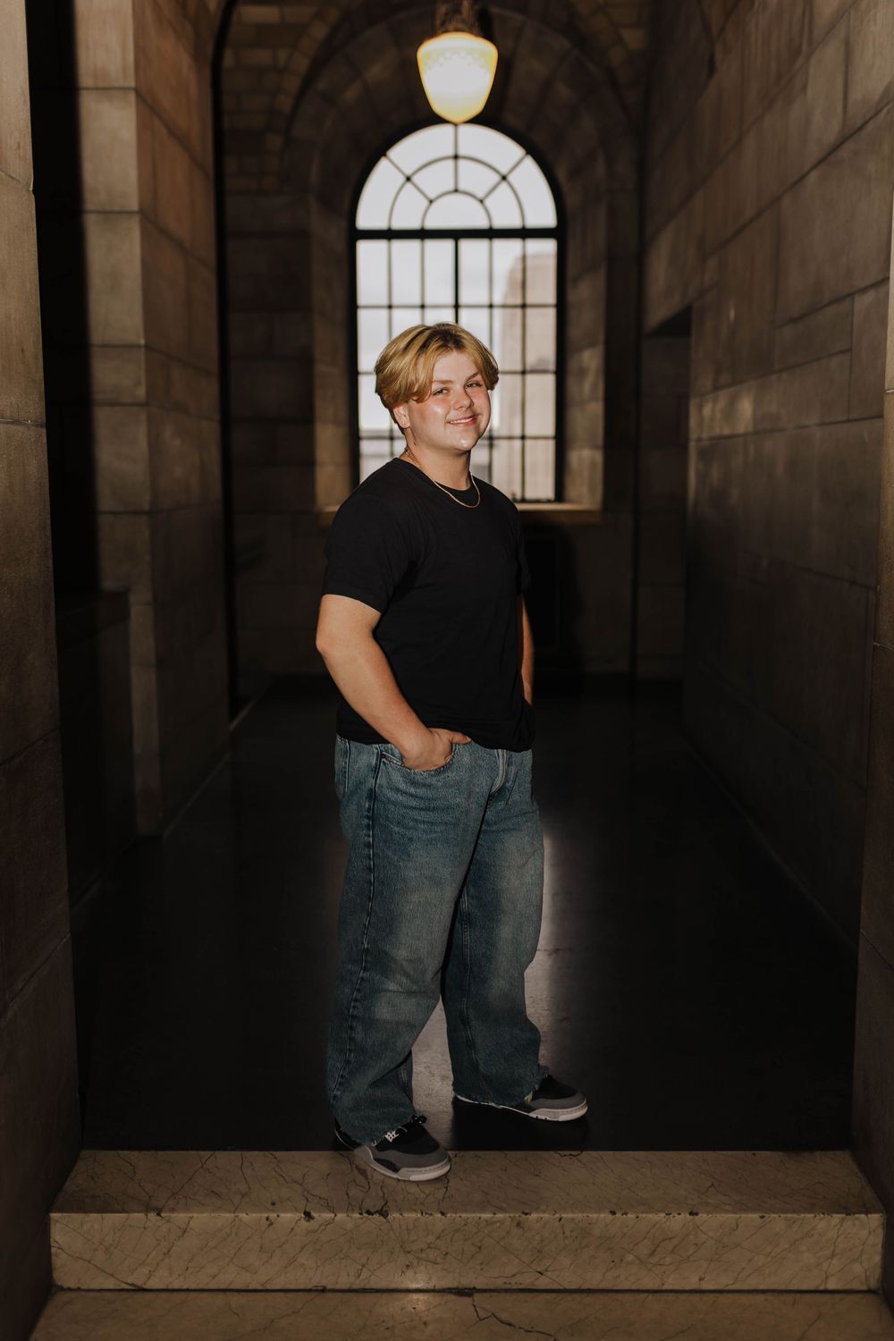 A person in a black t-shirt and jeans stands in a dark hallway with an arched window and stone walls.