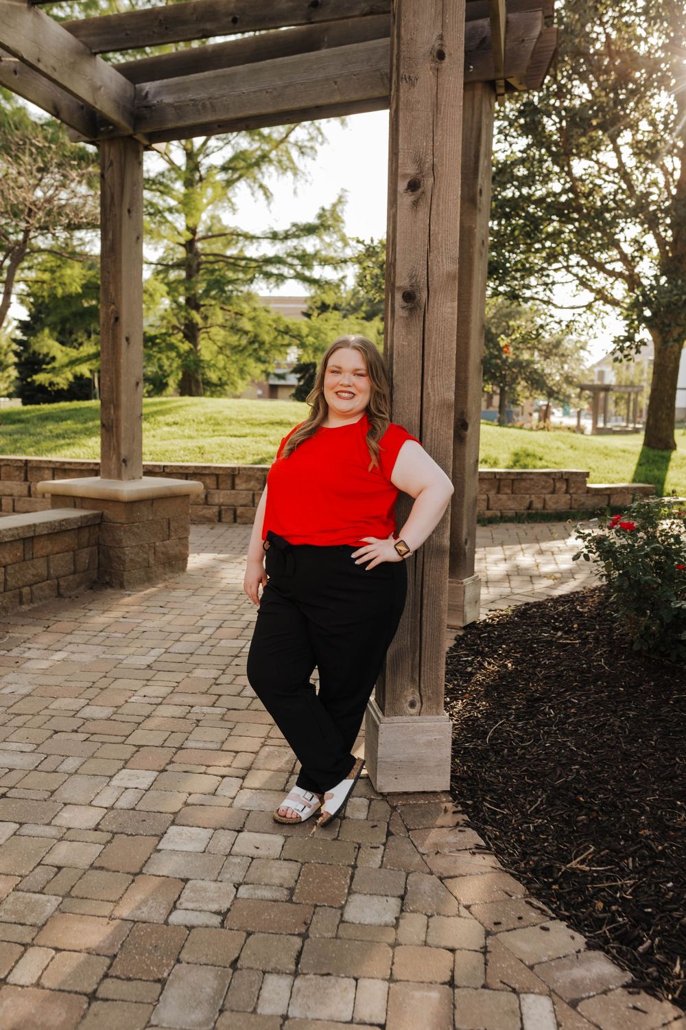 A person in a red top and black pants poses on stone pavers under a wooden pergola in an outdoor garden setting.