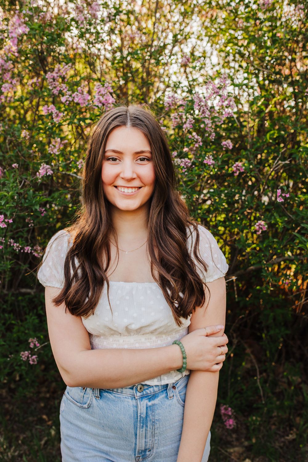 Portrait against flowering bushes showing a person in a white top and denim skirt during golden hour.