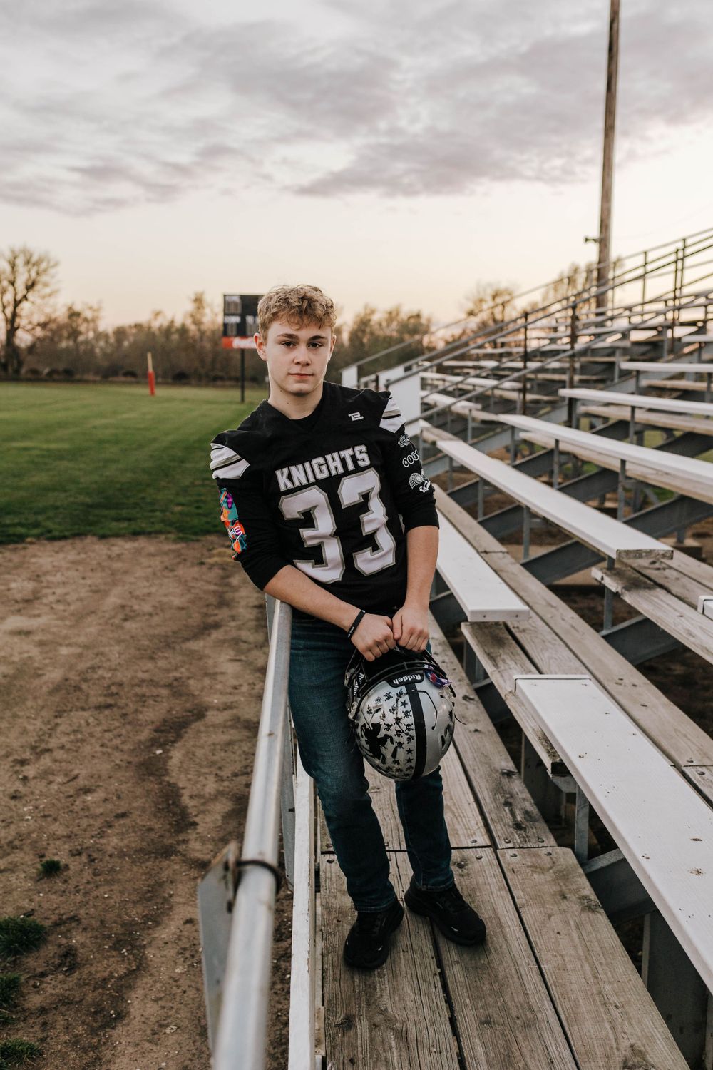 Football player in Knights jersey poses on bleachers at sunset holding helmet.